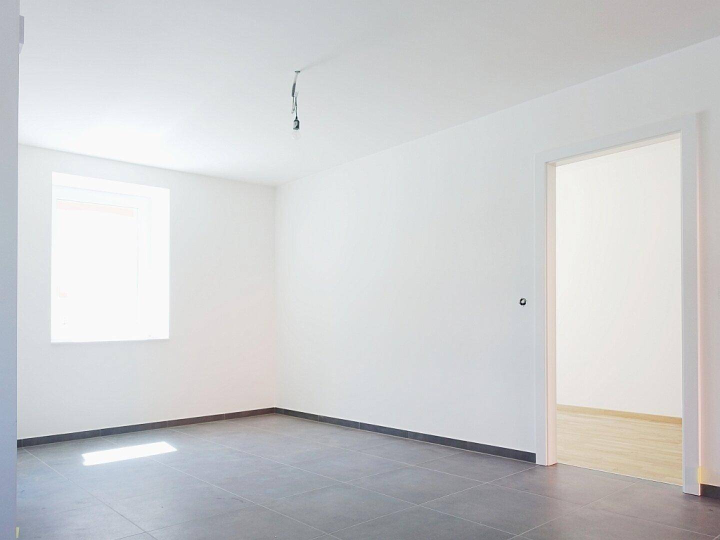 Geräumiger Raum mit grauen Fliesen und einem Fenster, ideal für flexible Nutzung in einem Neubau.