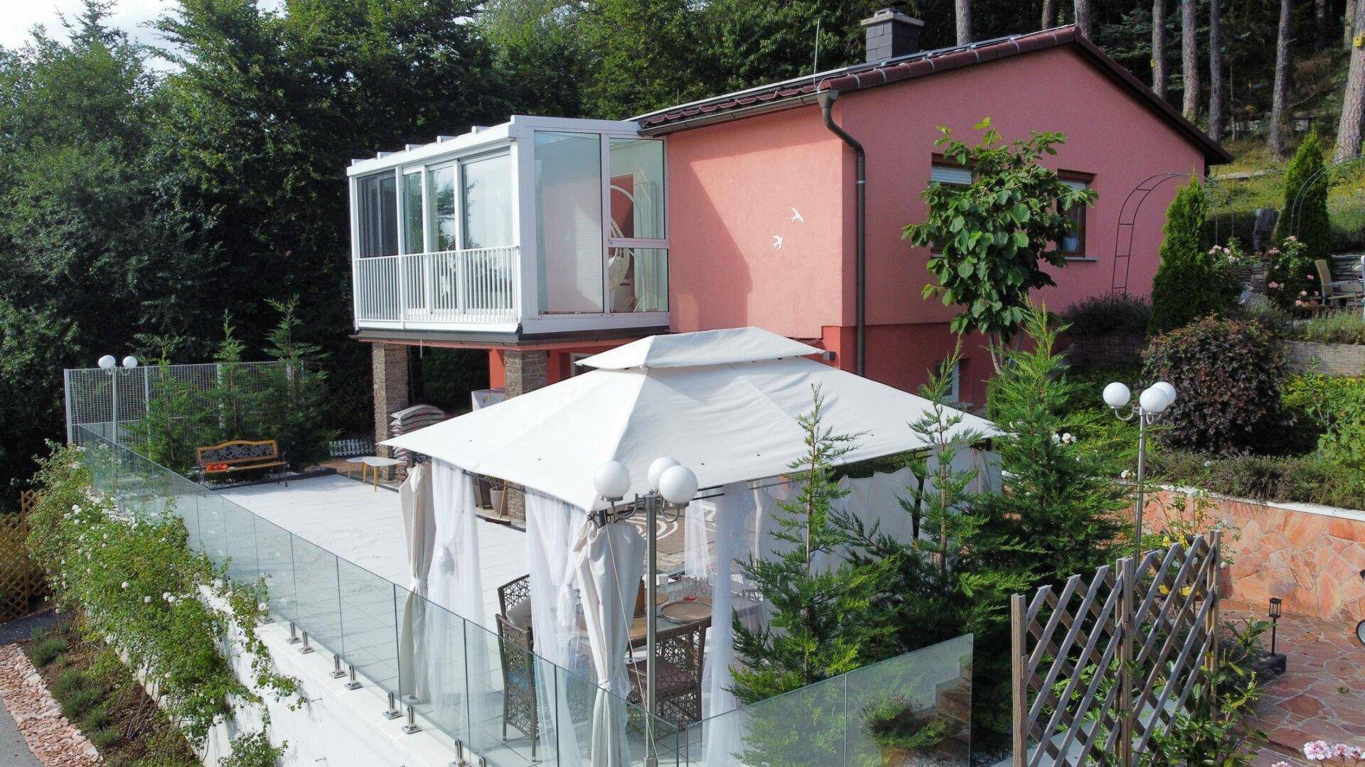 Blick auf die Rückseite des Hauses mit großer Terrasse, einem Pavillon und einem verglasten Wintergarten.