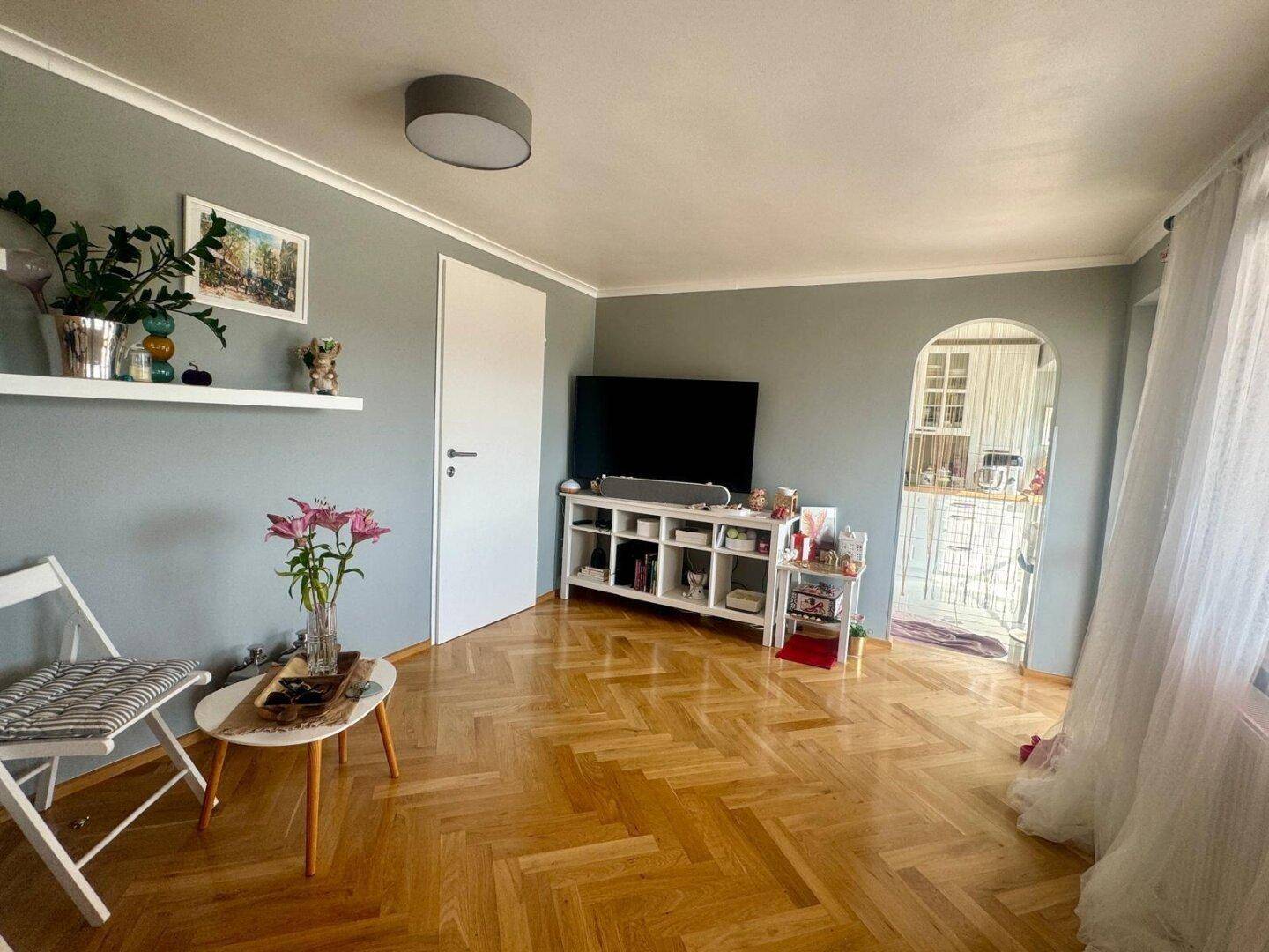 Gemütliches Wohnzimmer mit Fischgrätparkett, hellen Wänden und Blick in den Küchenbereich.