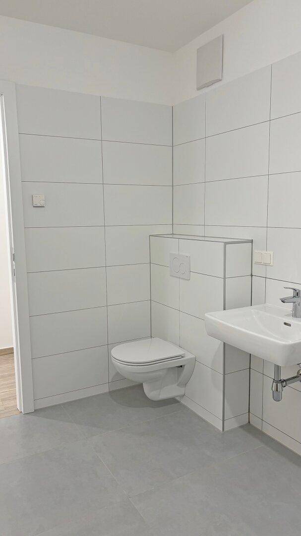 Modernes Badezimmer mit weißen Fliesen, grauen Bodenfliesen, wandhängendem WC und Waschbecken.