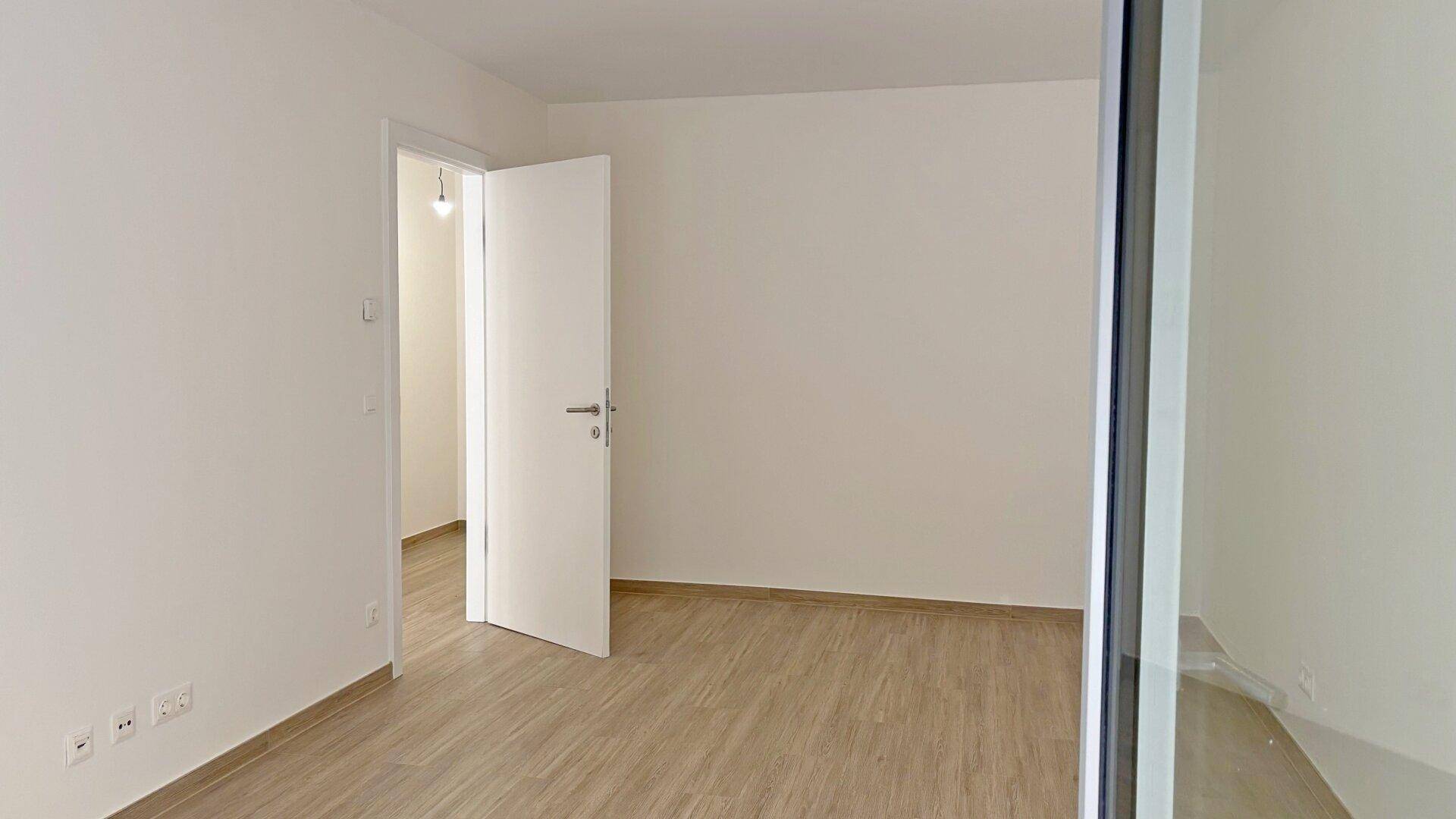 Heller, unmöblierter Raum mit Holzboden und offener Tür, die den Blick in einen weiteren Bereich freigibt.
