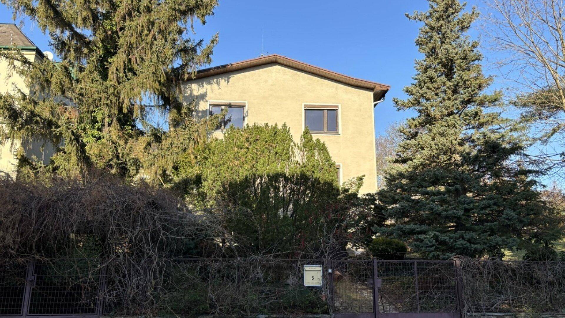 Vorderansicht des Hauses hinter einem Zaun und üppiger Bepflanzung bei Sonnenschein.