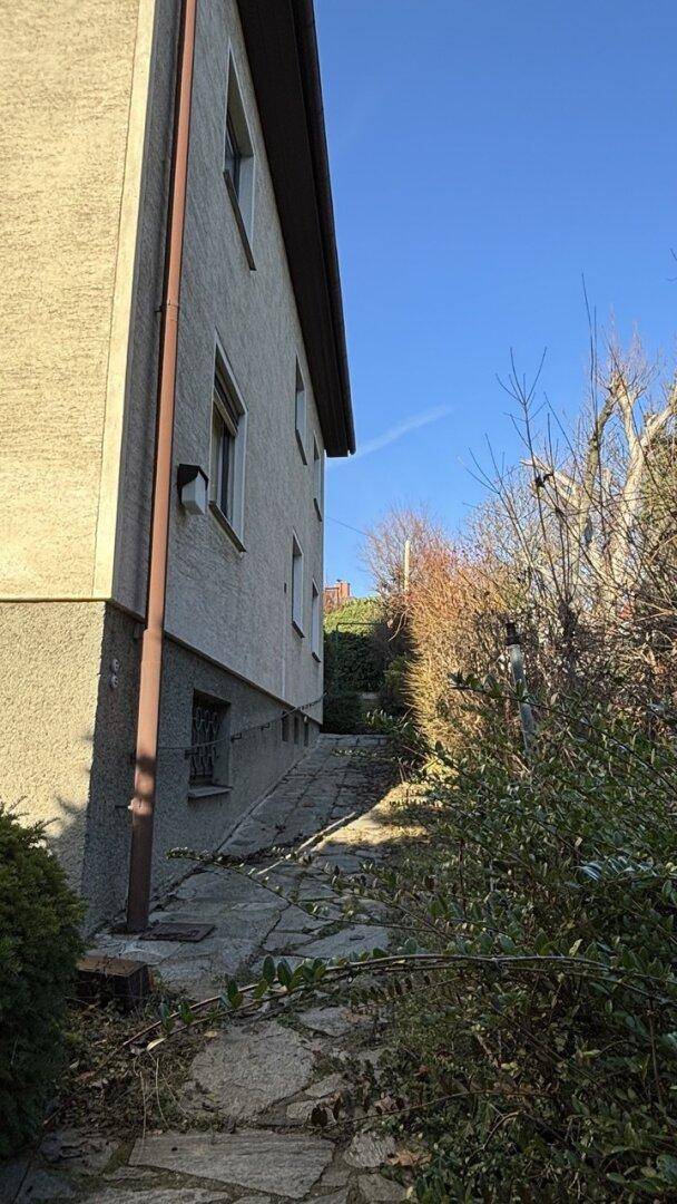 Seitenansicht des Hauses mit einem schmalen Weg entlang der Fassade und umgebender Vegetation.