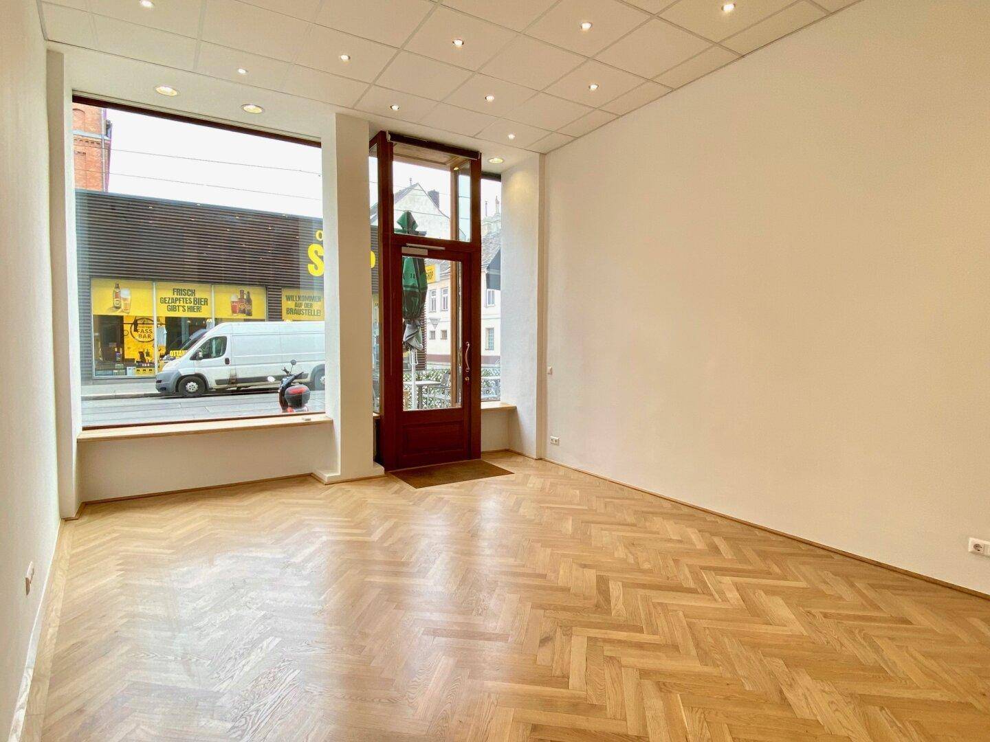 Heller Geschäftsraum mit Fischgrätparkett und großen Schaufenstern zur Straße, ideal für Ladenlokal.