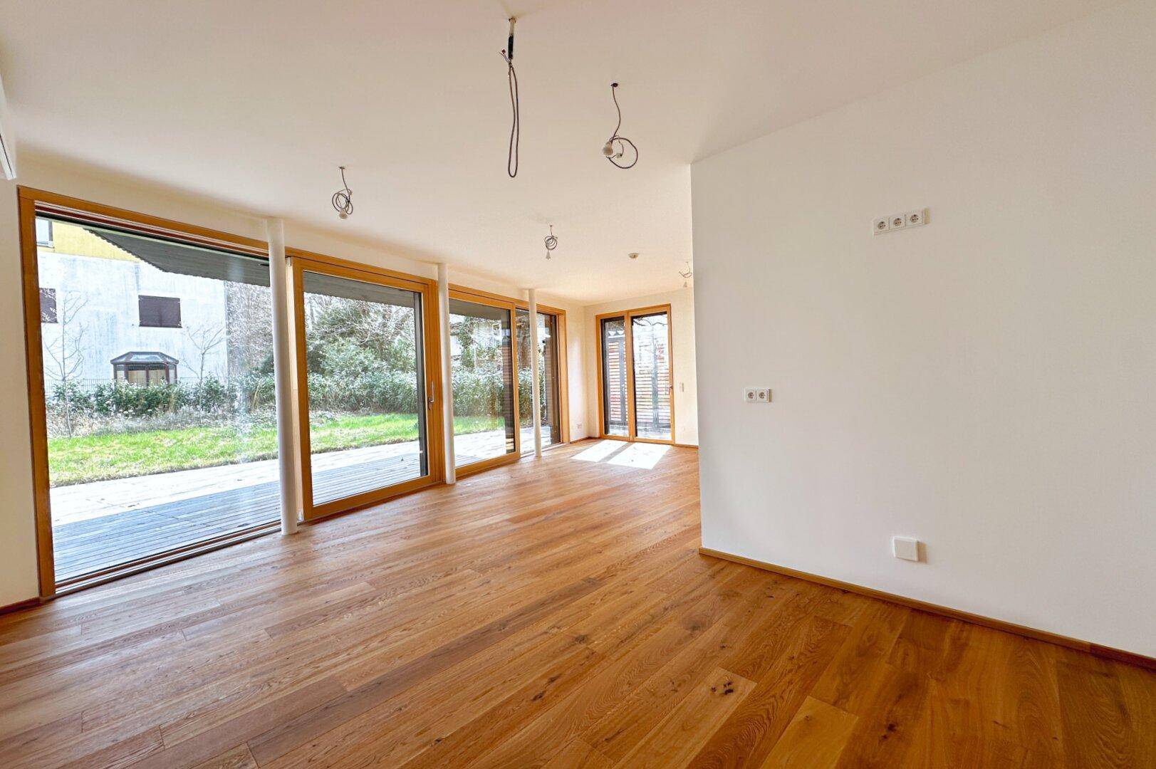 Helles, leeres Wohnzimmer mit Holzboden und großen Fenstern zum Garten.