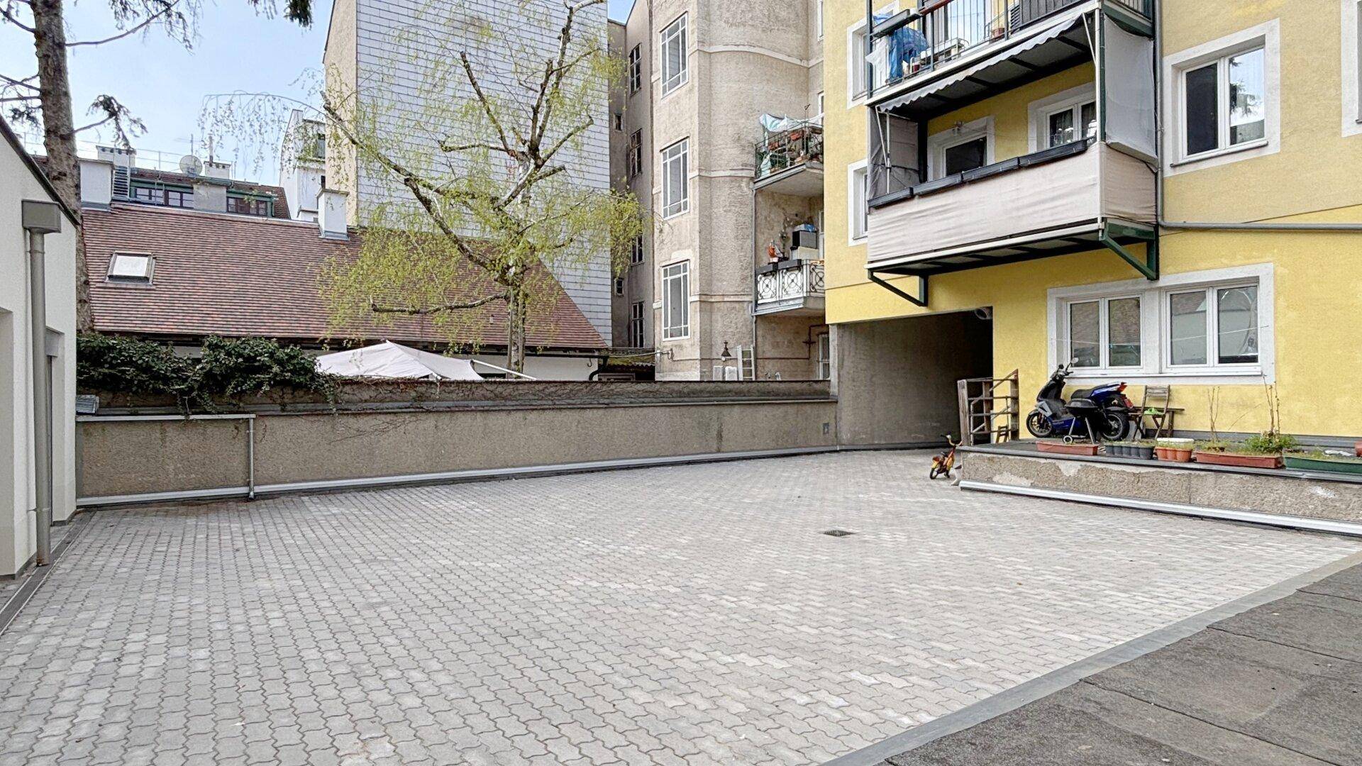 Gepflasterter Innenhof mit Parkmöglichkeiten, umgeben von Gebäuden, darunter ein älteres mit Balkonen.