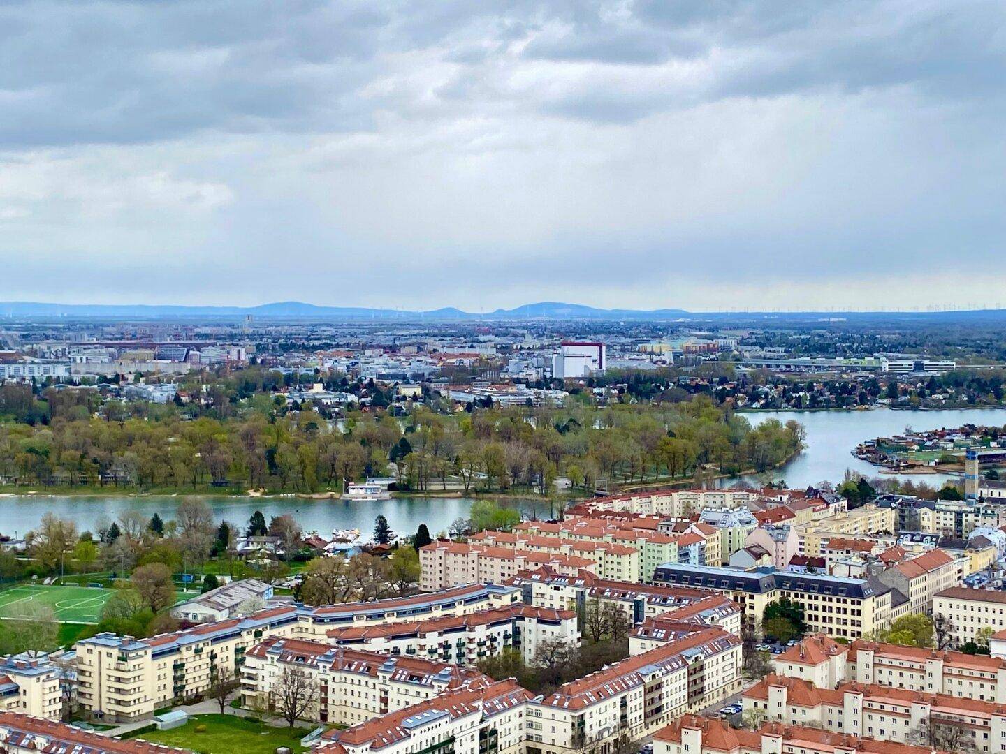 Weitläufiger Blick über die Stadt und die Donau mit grünen Inseln unter einem bewölkten Himmel.