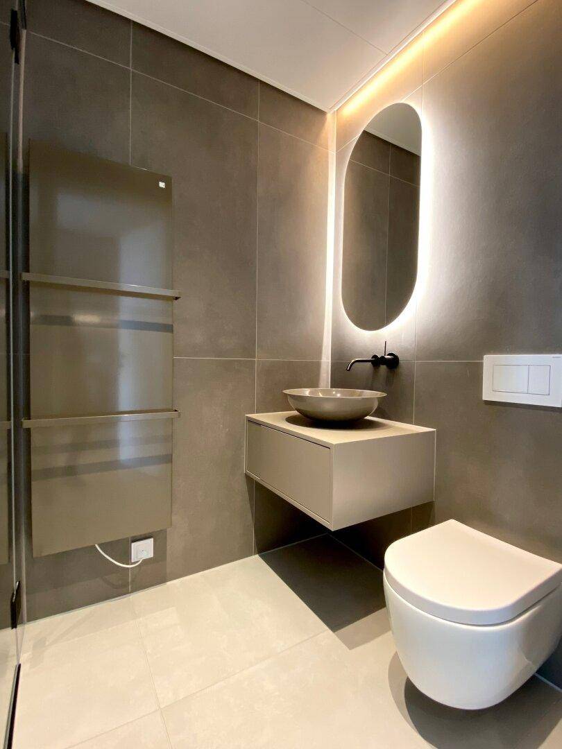 Modernes Badezimmer mit grauen Fliesen, schwebendem Waschtisch und beleuchtetem Spiegel.