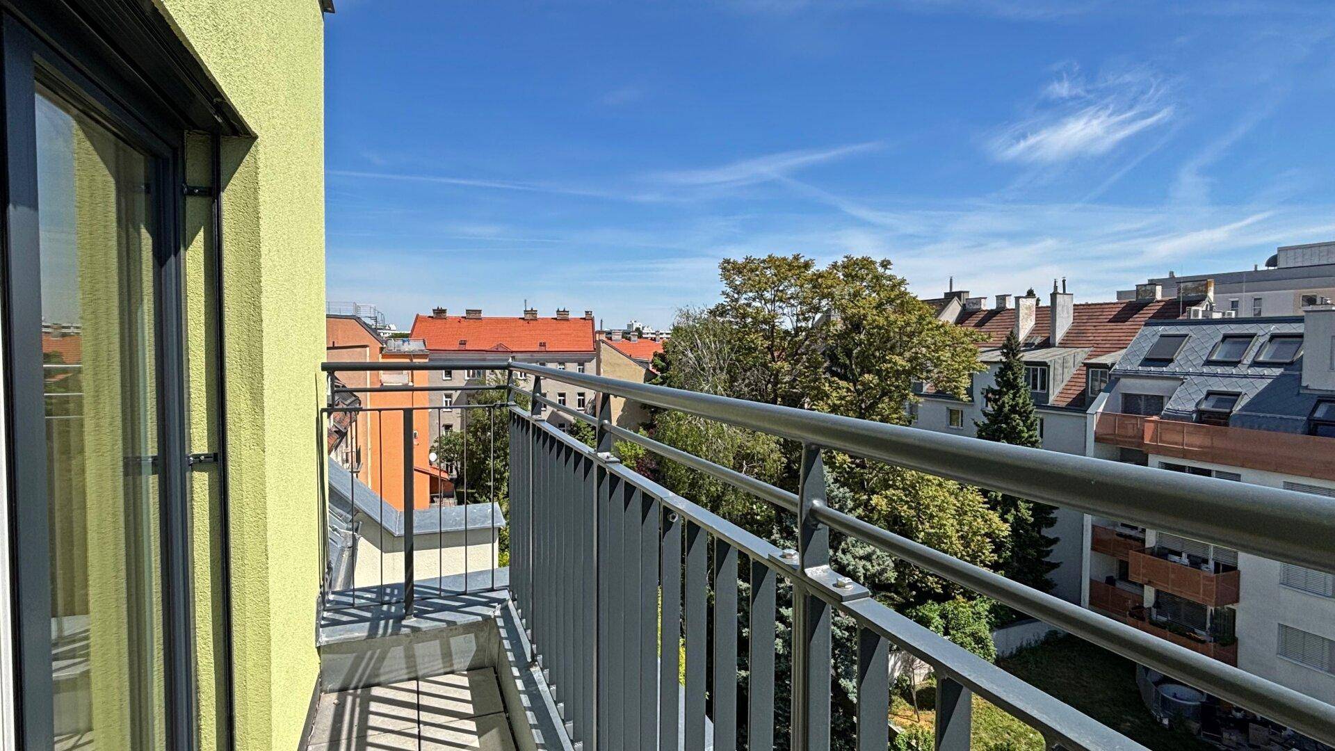 Balkon mit weitem Blick über die Dächer der Stadt und grüne Bäume unter blauem Himmel.
