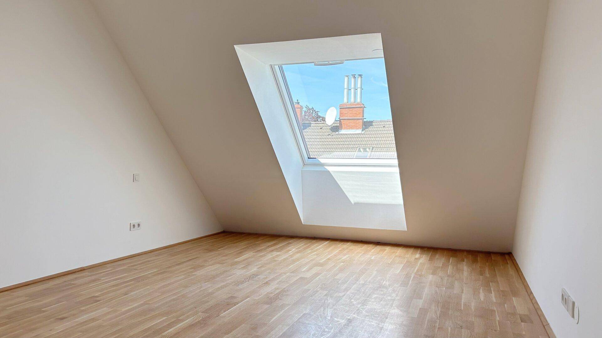 Dachzimmer mit schräger Decke, Holzboden und großem Dachfenster für viel Tageslicht.