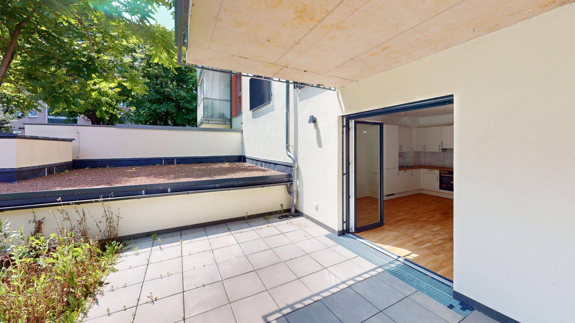 Sonniger Terrassenbereich mit Fliesenboden und direktem Zugang zum hellen Wohnraum mit Küchenzeile.