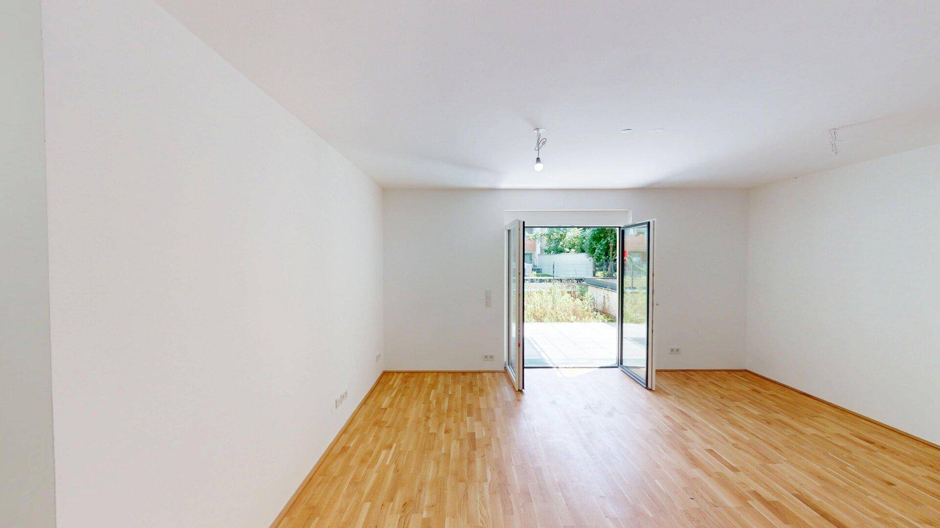 Geräumiger, leerer Wohnraum mit hellem Parkettboden und großen Fenstern zum Außenbereich.