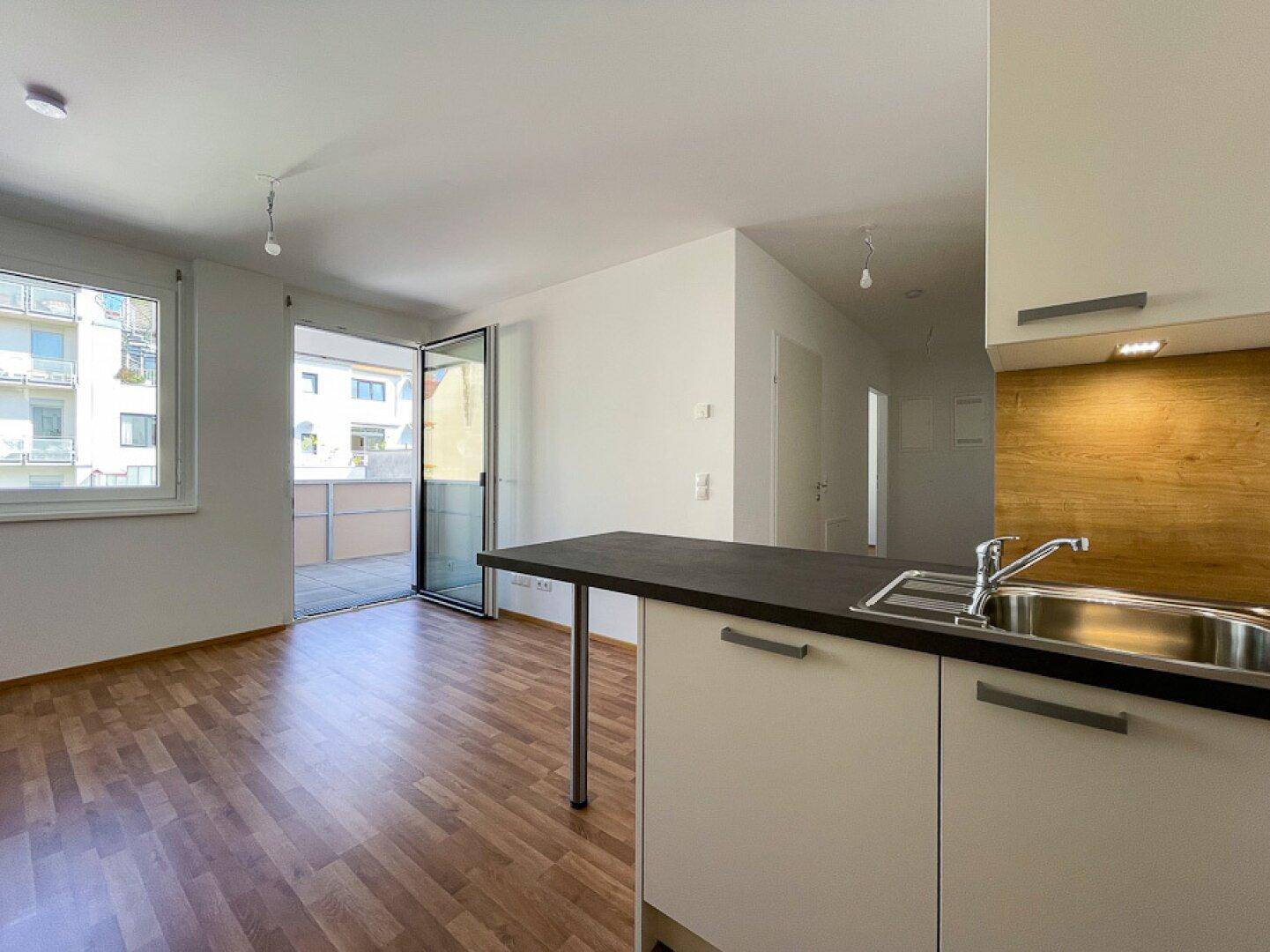 Offener Wohnbereich mit Laminatboden, Küchenzeile und Zugang zu einem Balkon.