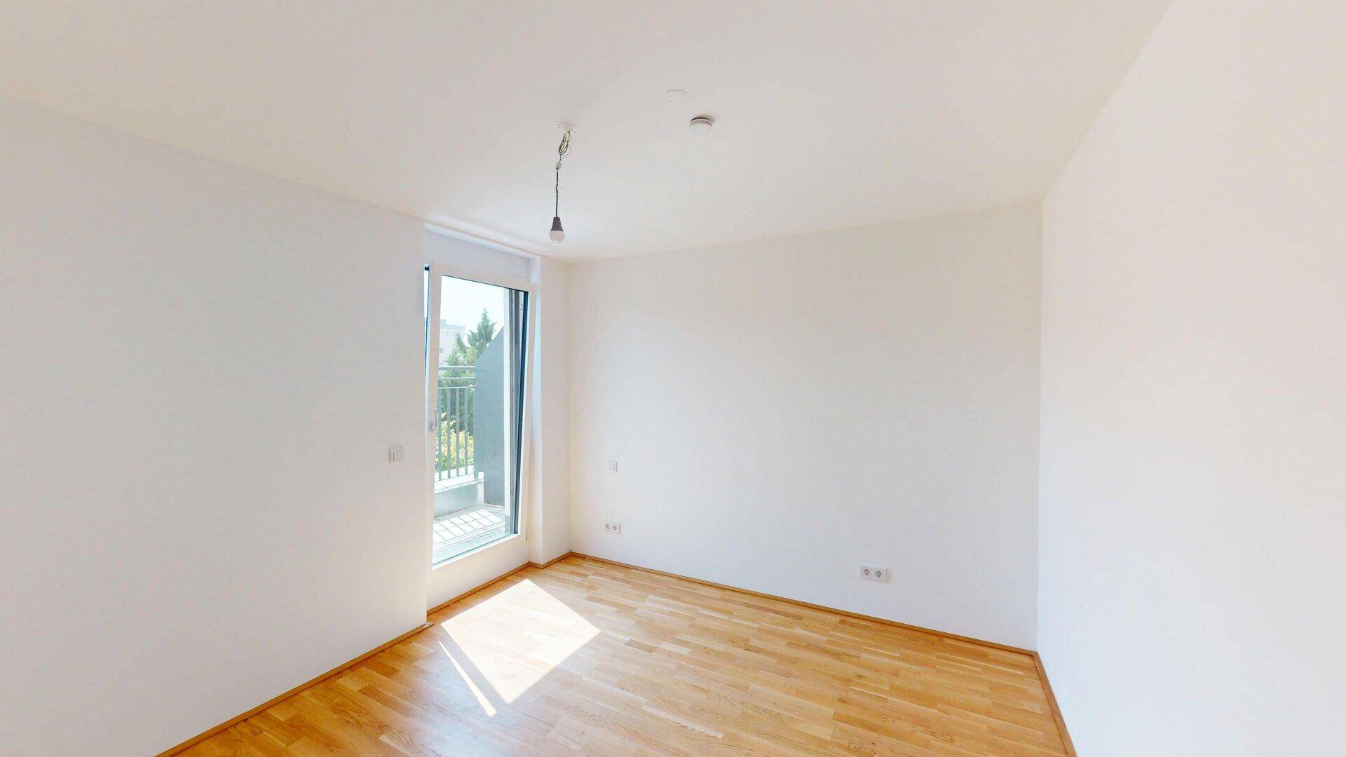 Unmöblierter Raum mit Holzboden und Balkonzugang, der viel Sonnenlicht empfängt.