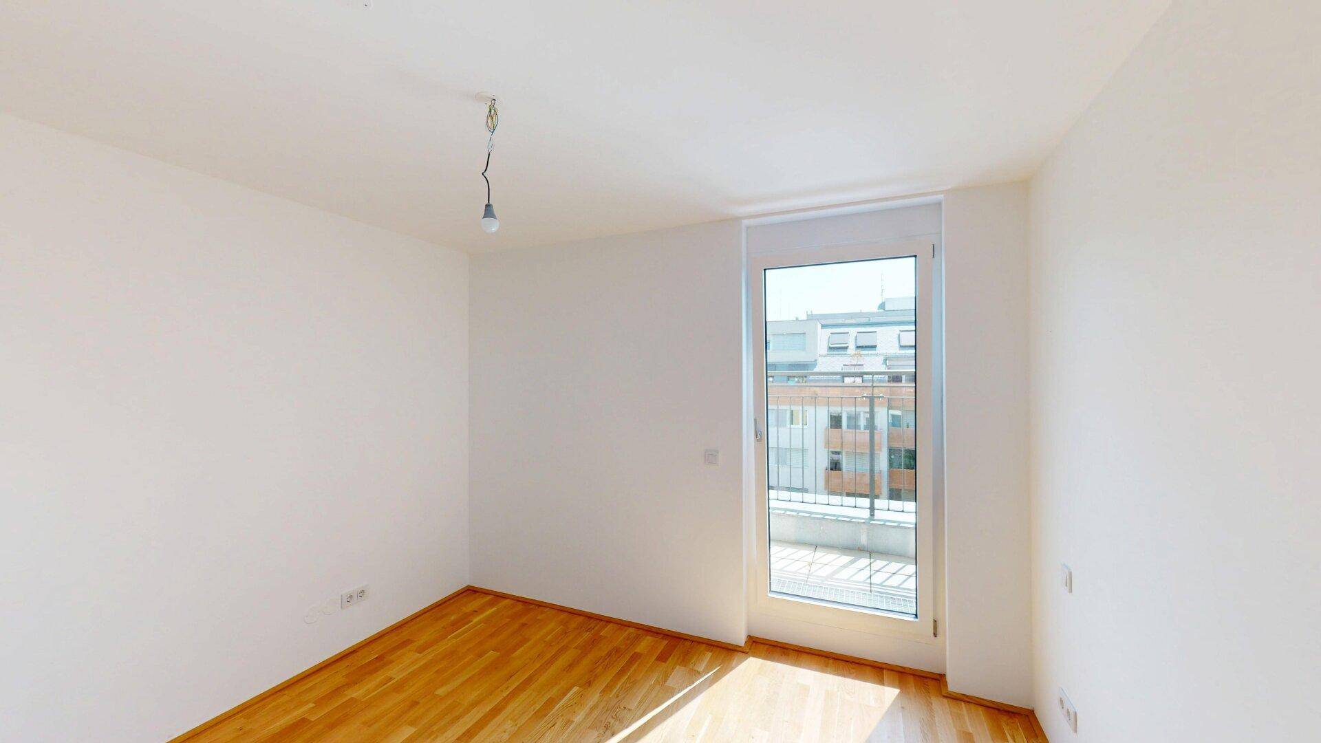 Leerer Raum mit Parkettboden und Zugang zu einem Balkon, hell durch große Fenster.