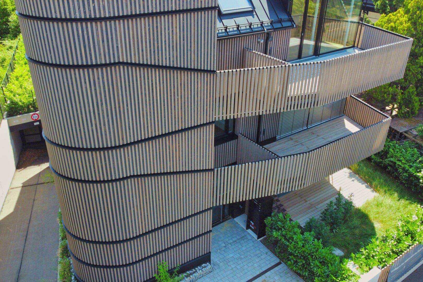 Luftaufnahme eines modernen Gebäudes mit geschwungener Holzlamellenfassade und mehreren Balkonen.