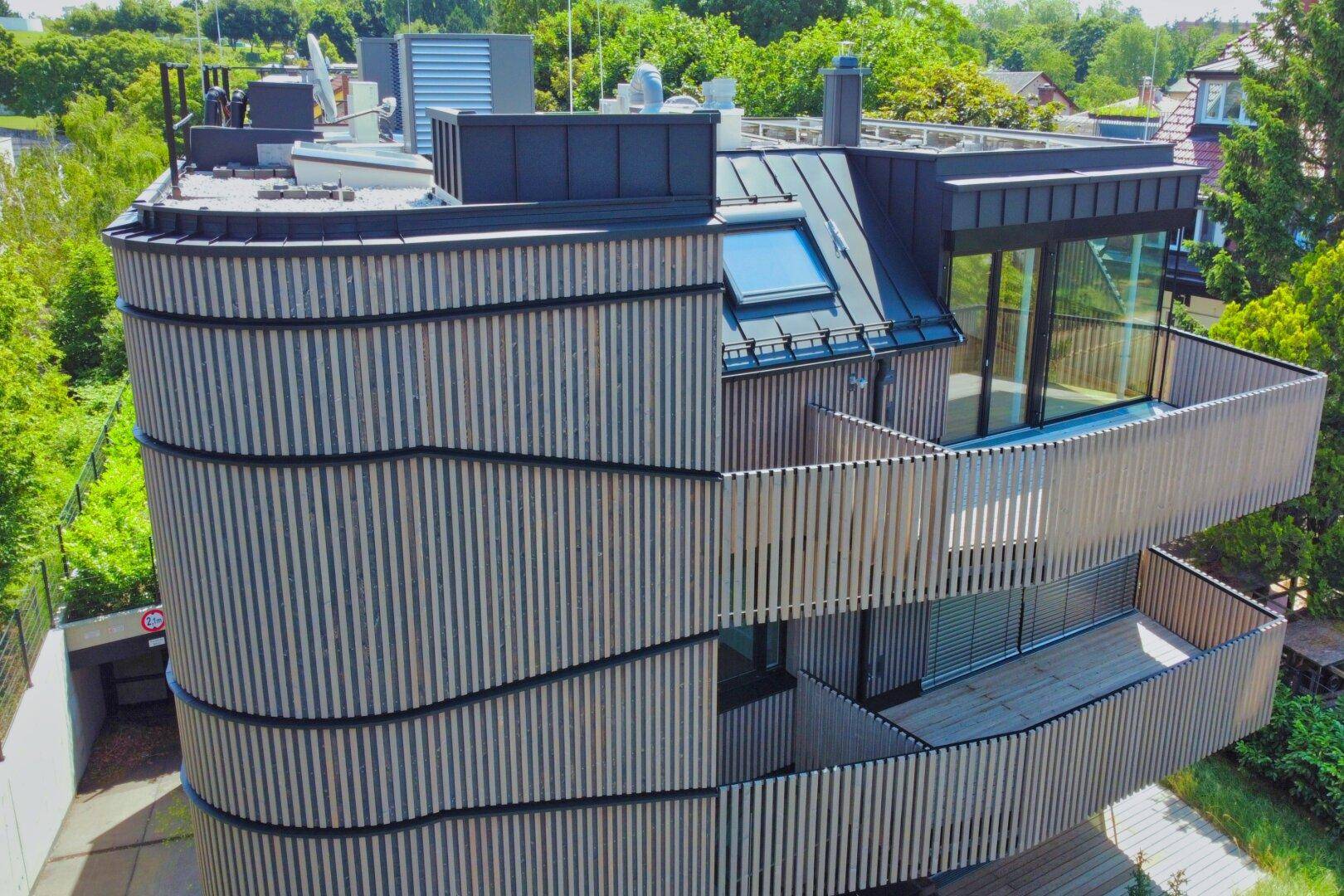 Luftaufnahme des modernen Gebäudes mit geschwungener Holzfassade und Dachfenster.