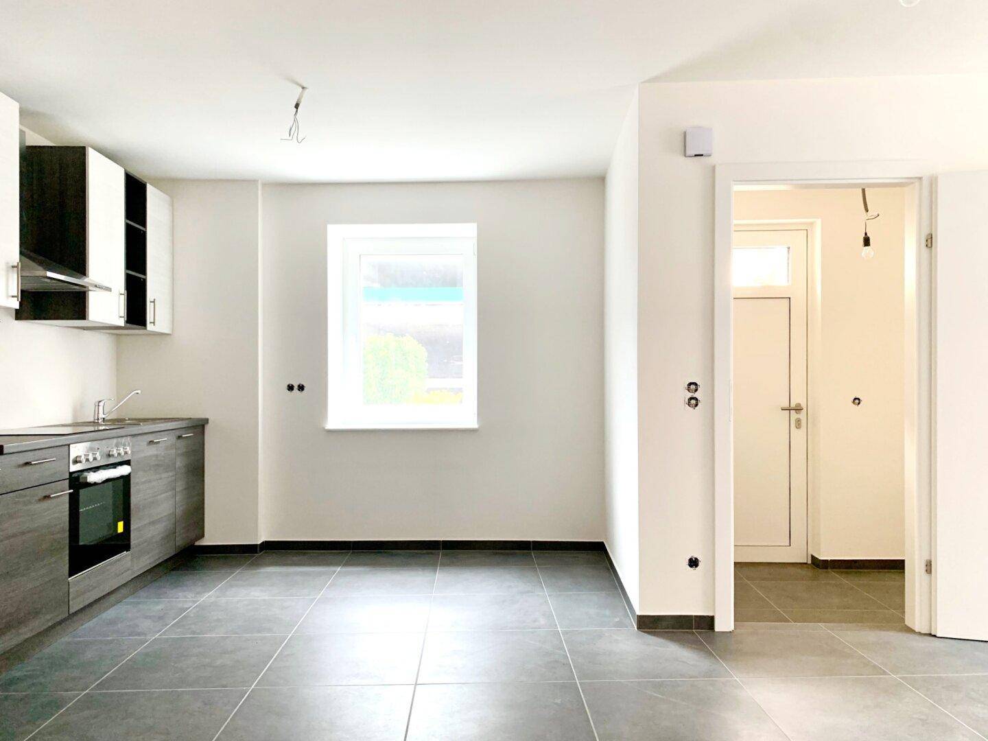 Helle Küche mit grauen und weißen Schränken, Fenster und Zugang zu einem weiteren Raum mit Fliesenboden.