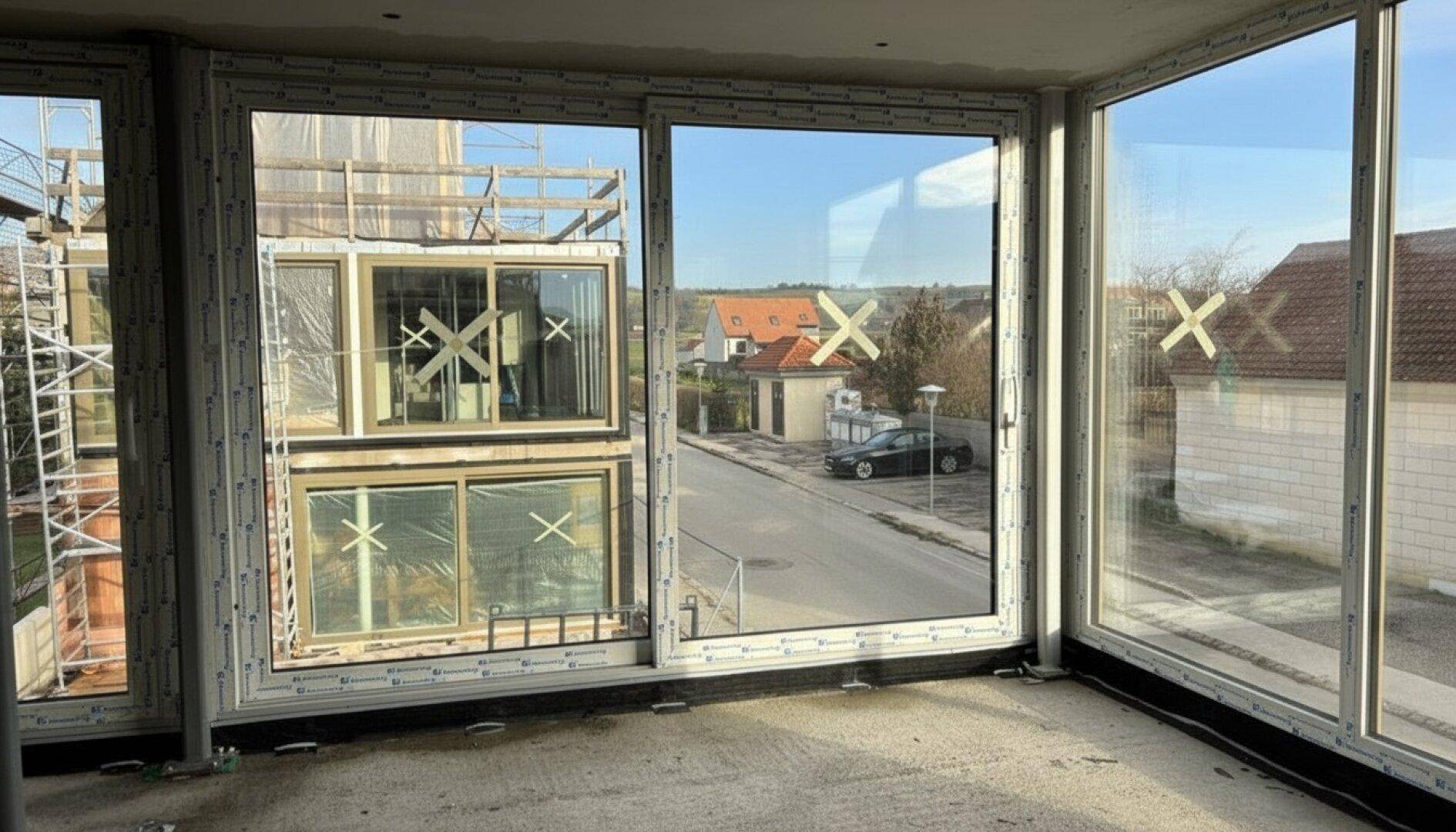 Innenraum im Rohbau mit großen Fensterfronten und Blick auf die Straße und Nachbargebäude.