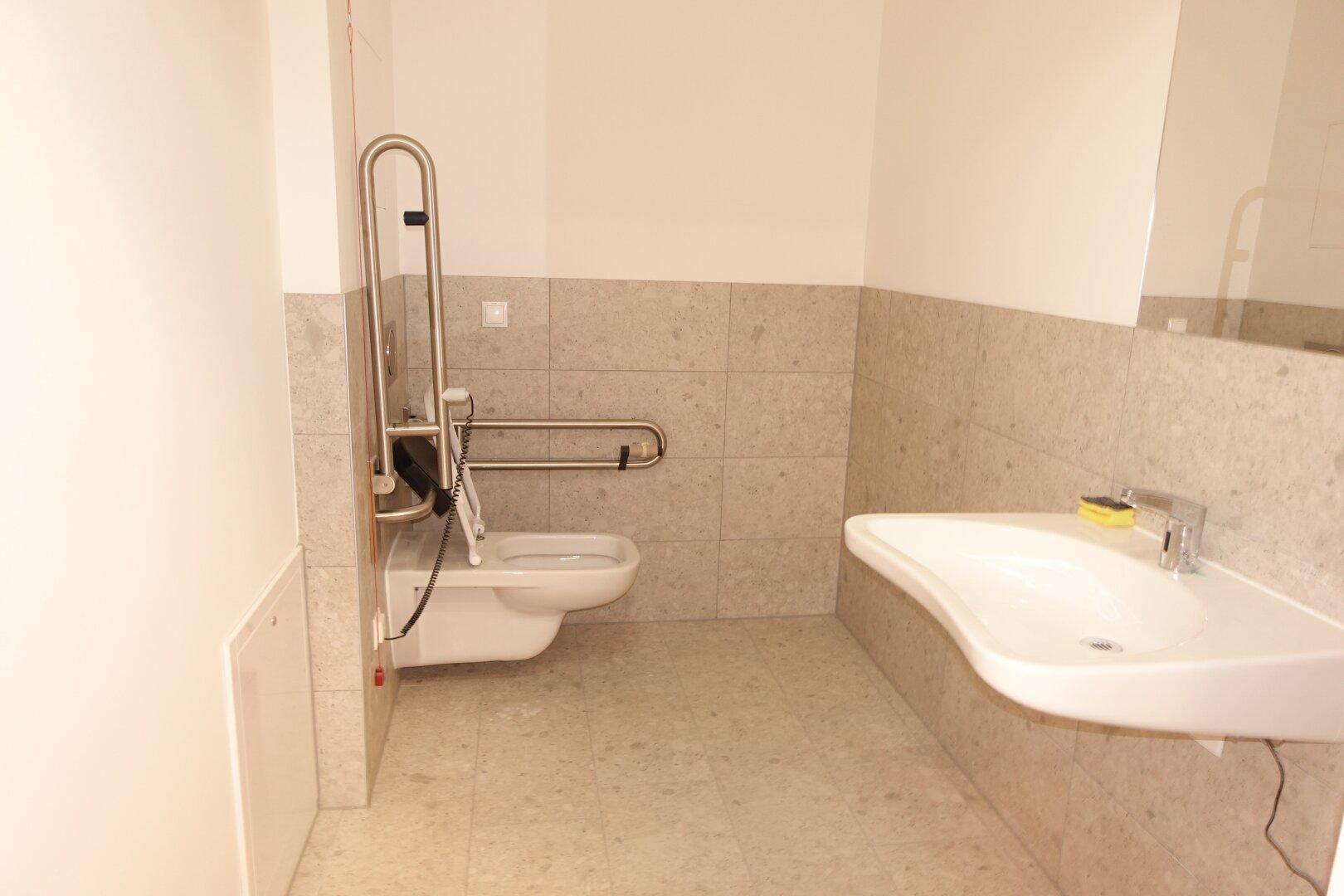 Barrierefreies Badezimmer mit Haltegriffen, wandhängender Toilette und modernem Waschbecken.