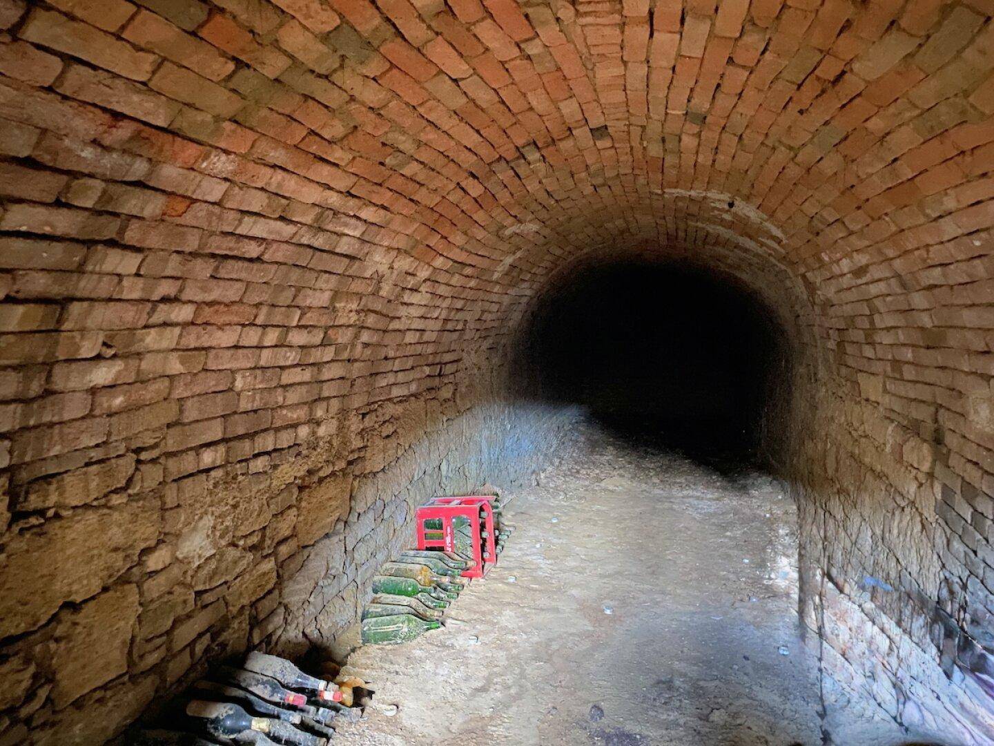 Alter Gewölbekeller mit Ziegelwänden und gelagerten Flaschen, der eine historische Atmosphäre ausstrahlt.