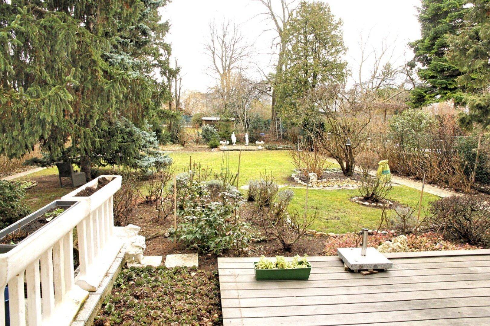 Gepflegter Garten mit großer Rasenfläche, umrandeten Beeten und altem Baumbestand, von oben betrachtet.