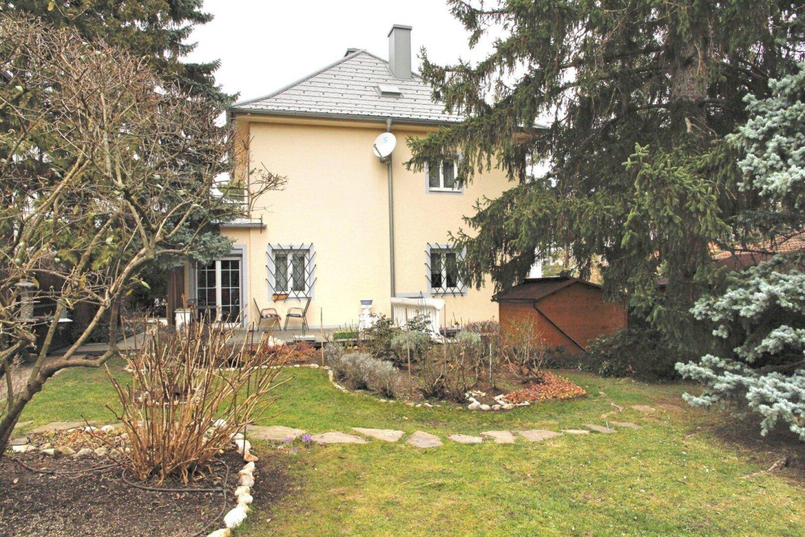 Rückansicht des Hauses mit Terrasse und weitläufigem Garten, umgeben von altem Baumbestand.