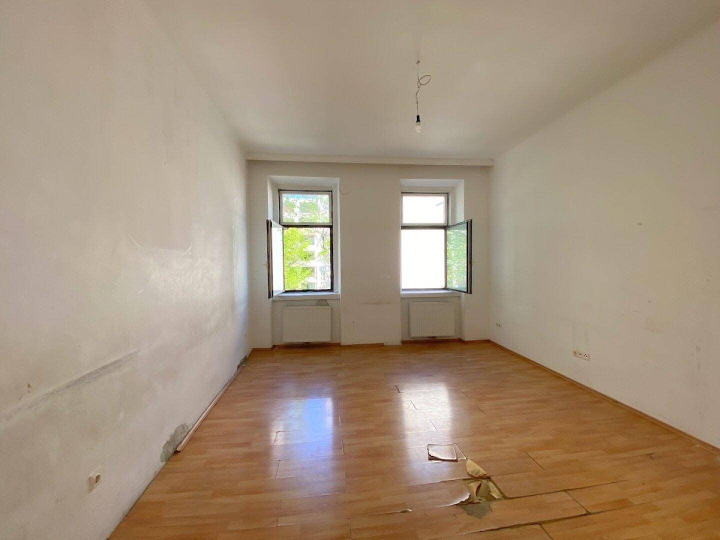 Heller, leerer Raum mit zwei Fenstern, Parkettboden und deutlichen Gebrauchsspuren, bereit für eine Sanierung.