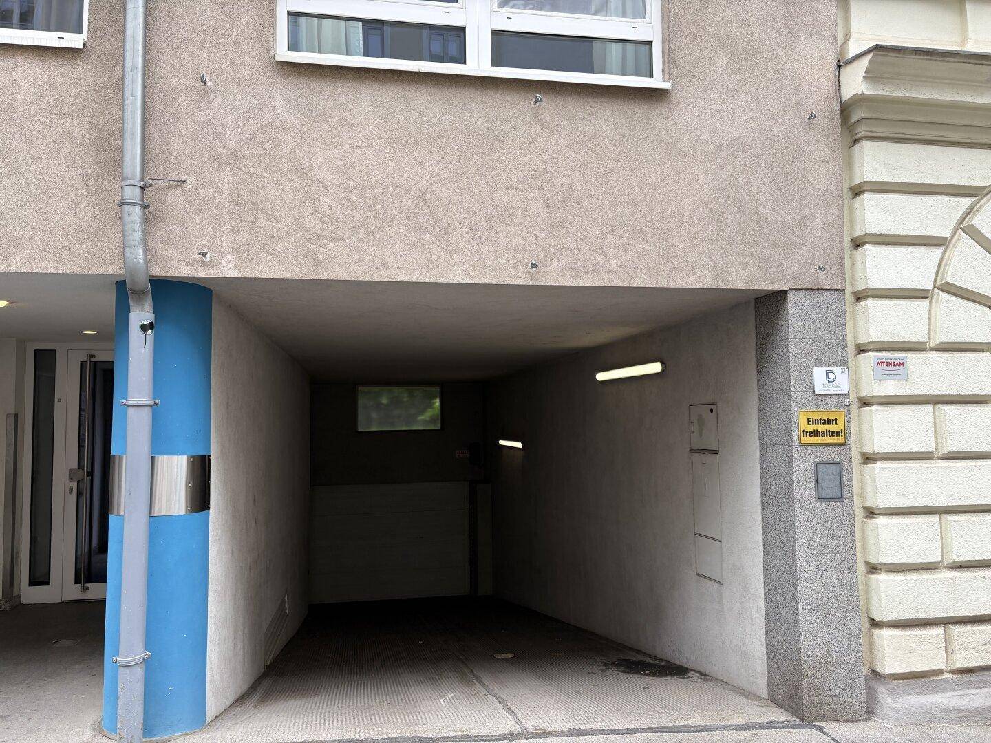 Einfahrt zu einer Tiefgarage mit blauer Säule und darüberliegender Gebäudefassade.