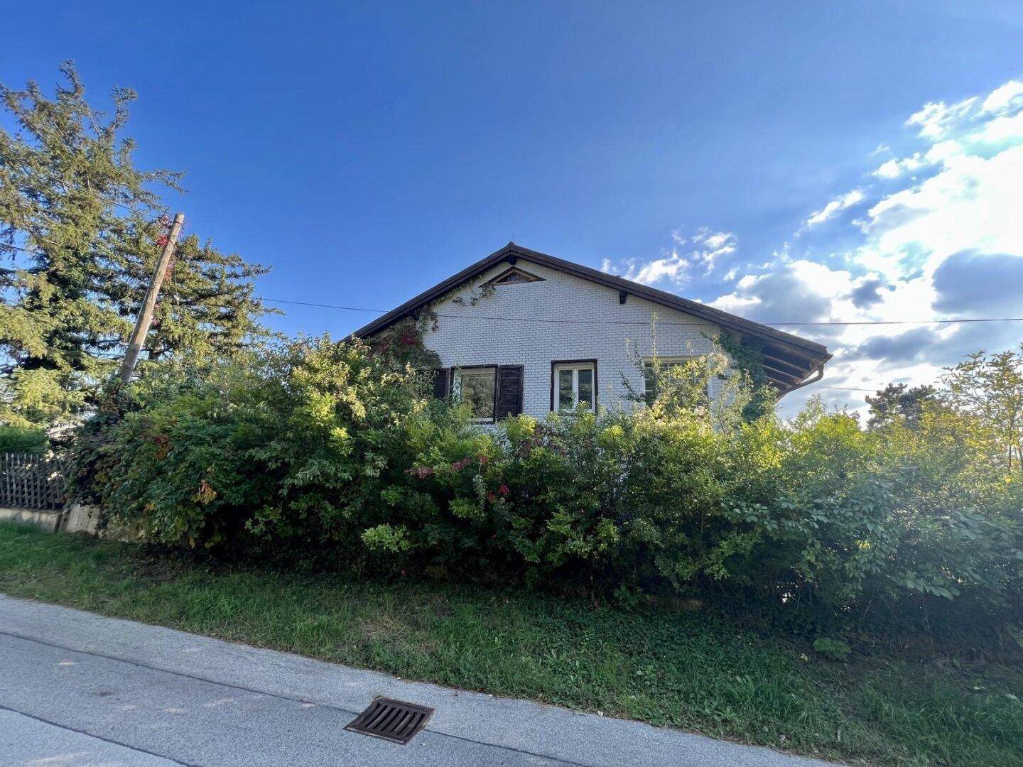 Älteres Wohnhaus mit Giebeldach, umgeben von üppiger Vegetation und Sträuchern unter blauem Himmel.