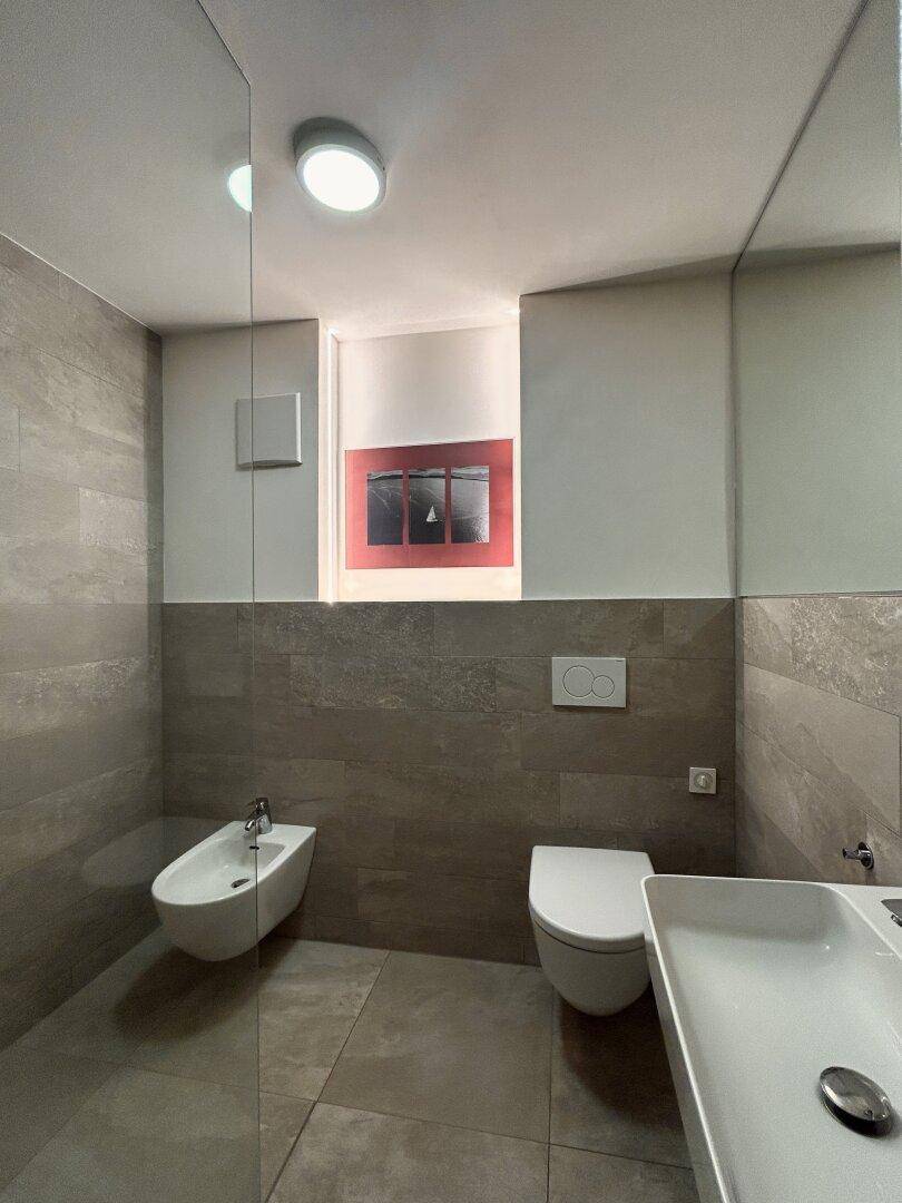 Modernes Badezimmer mit WC, Bidet und einer begehbaren Dusche, ausgestattet mit grauen Fliesen.