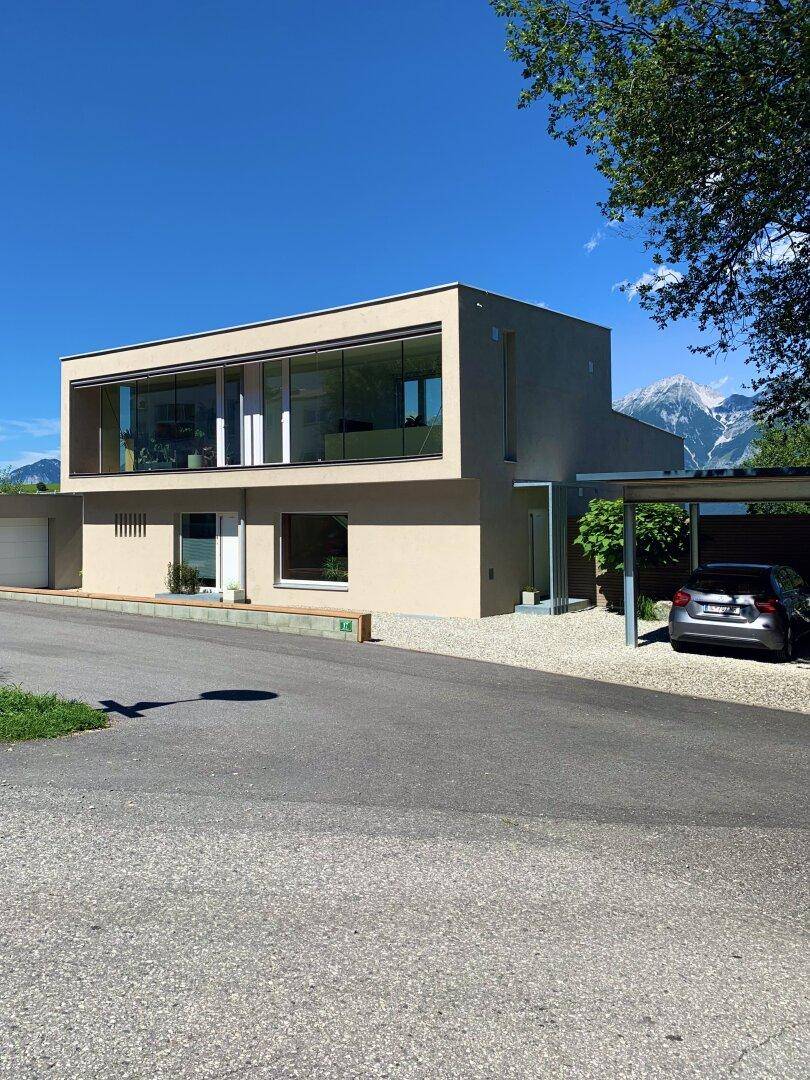 Modernes Wohnhaus mit Carport und Zufahrt, umgeben von grüner Landschaft und Bergen.