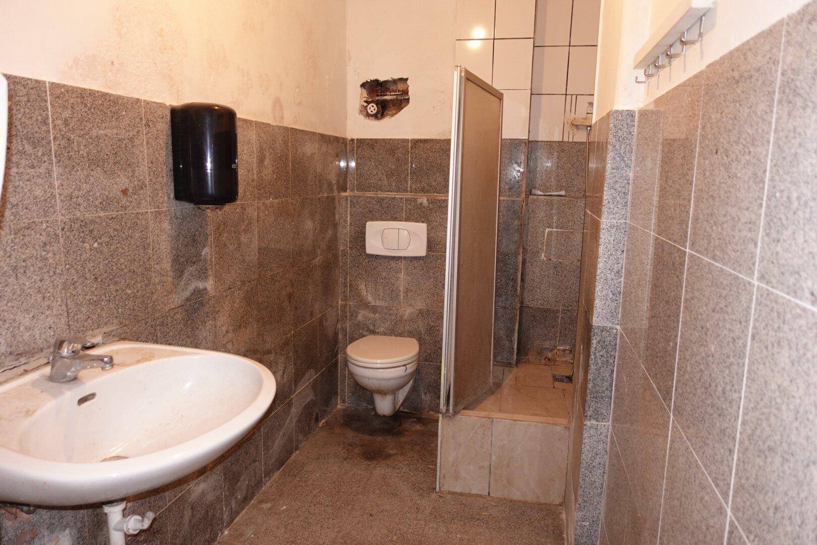 Badezimmer mit grauen Fliesen an Wänden und Boden, ausgestattet mit Waschbecken, Toilette und Duschbereich.