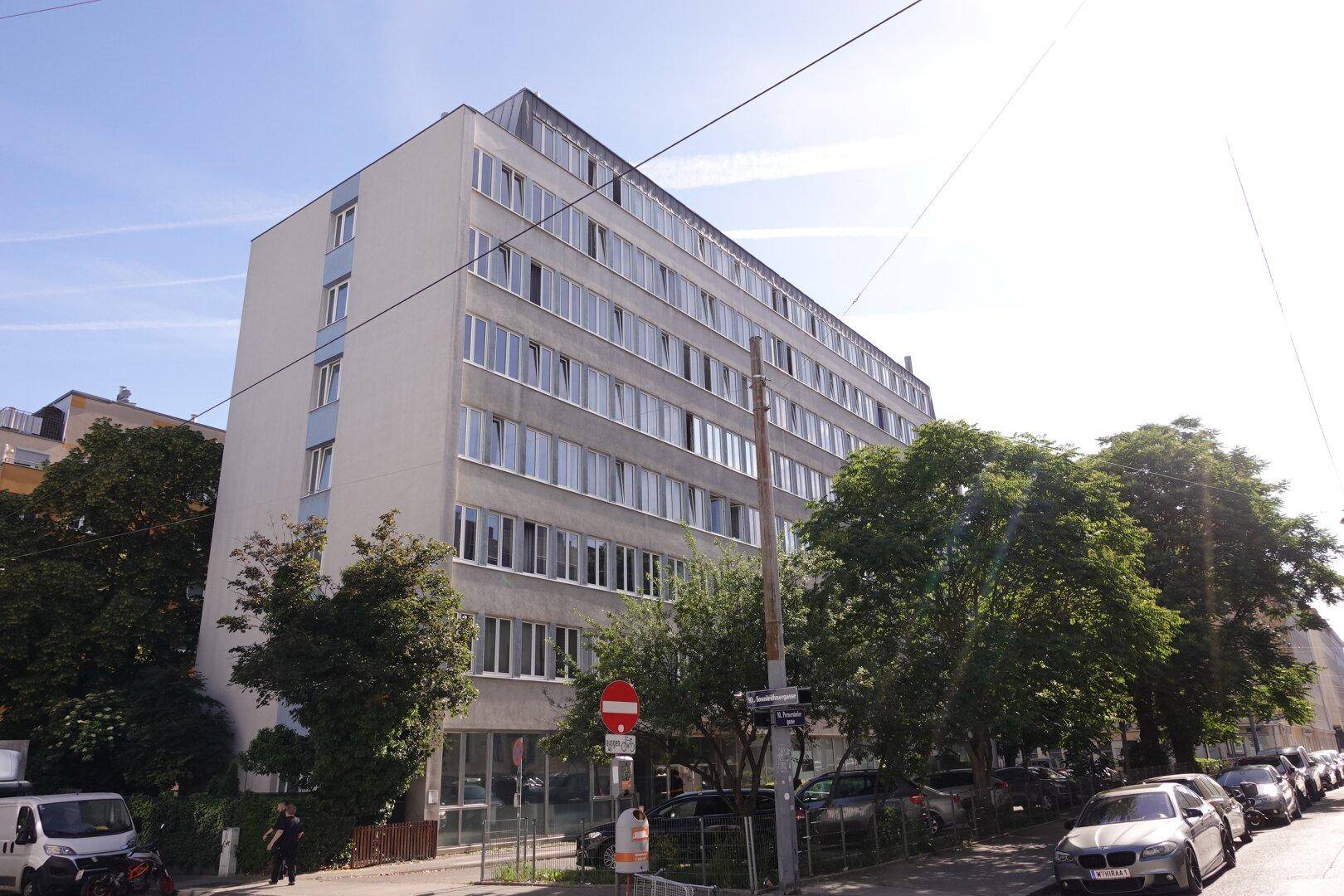 Mehrstöckiges Gebäude mit zahlreichen Fenstern und Bäumen entlang der Straße, in städtischer Umgebung.