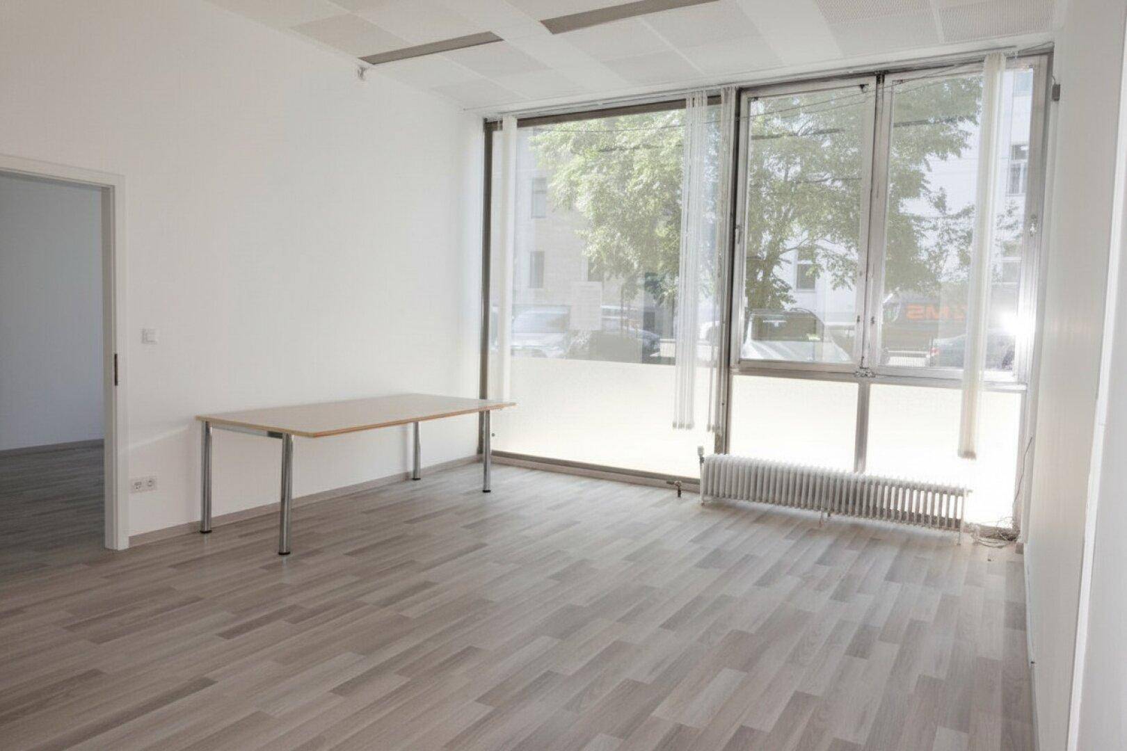 Heller Büroraum mit großen Fenstern, Laminatboden und einem einfachen Tisch, gut beleuchtet.