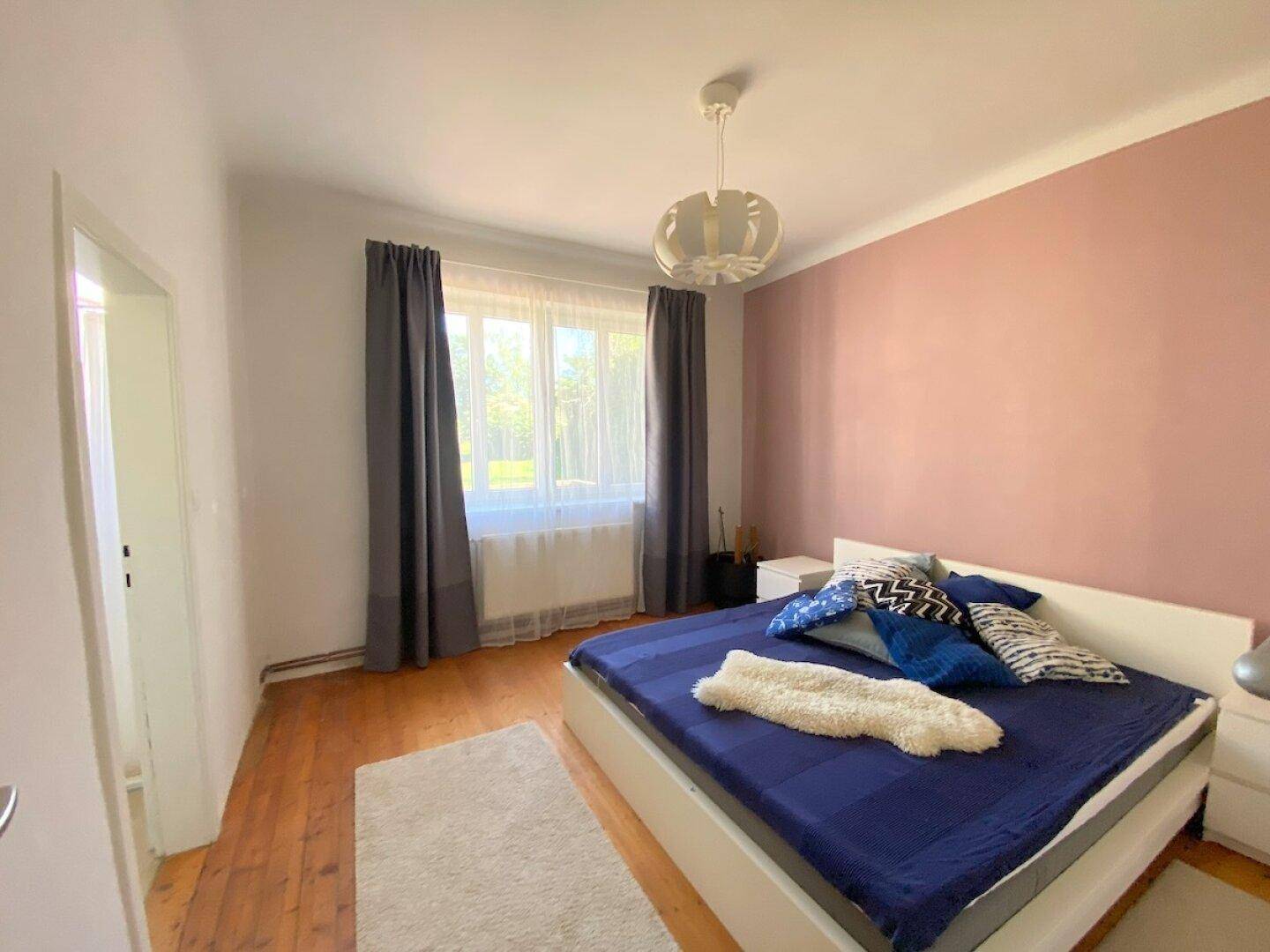 Schlafzimmer mit Holzboden, rosa Akzentwand und großem Fenster für Tageslicht.