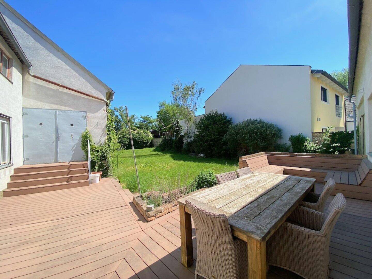 Große Holzterrasse mit Gartenmöbeln und angrenzender Rasenfläche unter blauem Himmel.