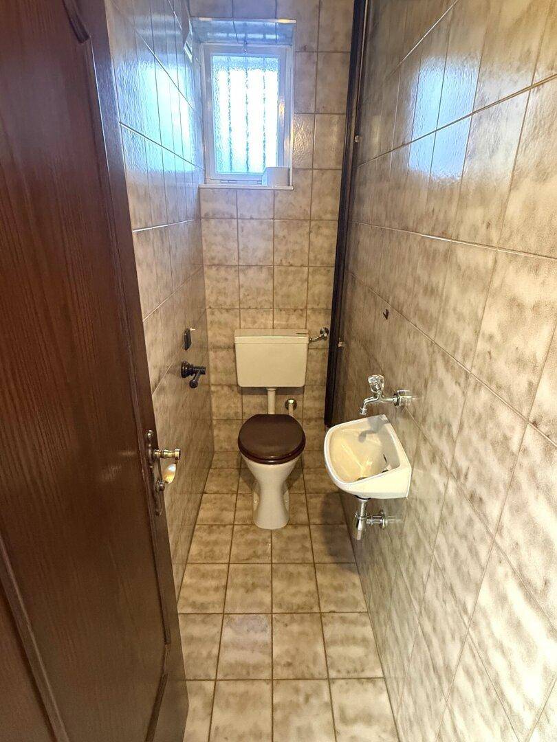 Separate Toilette mit beigen Fliesen, WC und kleinem Handwaschbecken.