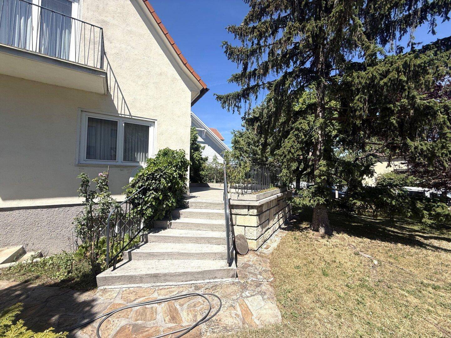 Seitlicher Blick auf das Haus mit Außentreppe, Balkon und umgebender Vegetation.