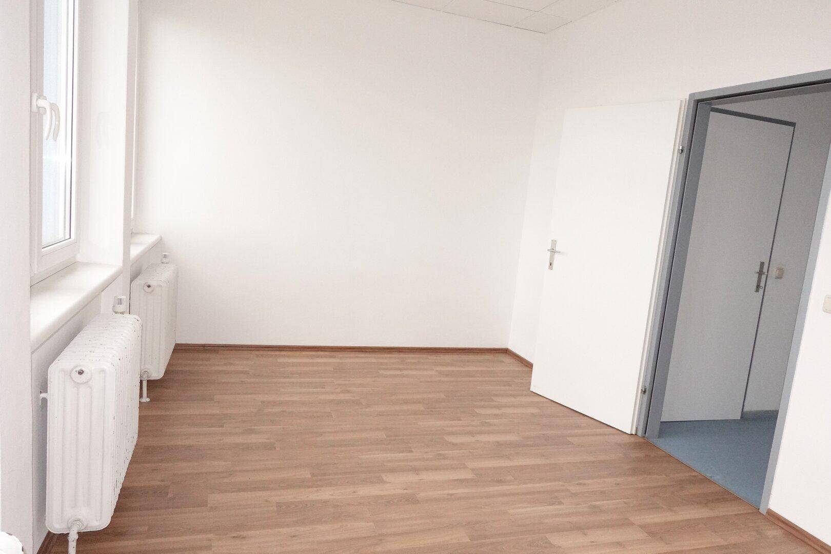 Leeres Zimmer mit Laminatboden und Fenster, das Zugang zu einem weiteren Raum bietet.