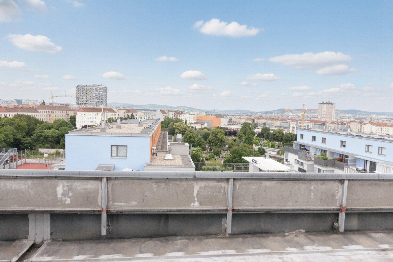 Weitläufiger Blick über die Dächer der Stadt mit blauen Gebäuden und bewaldeten Hügeln im Hintergrund.