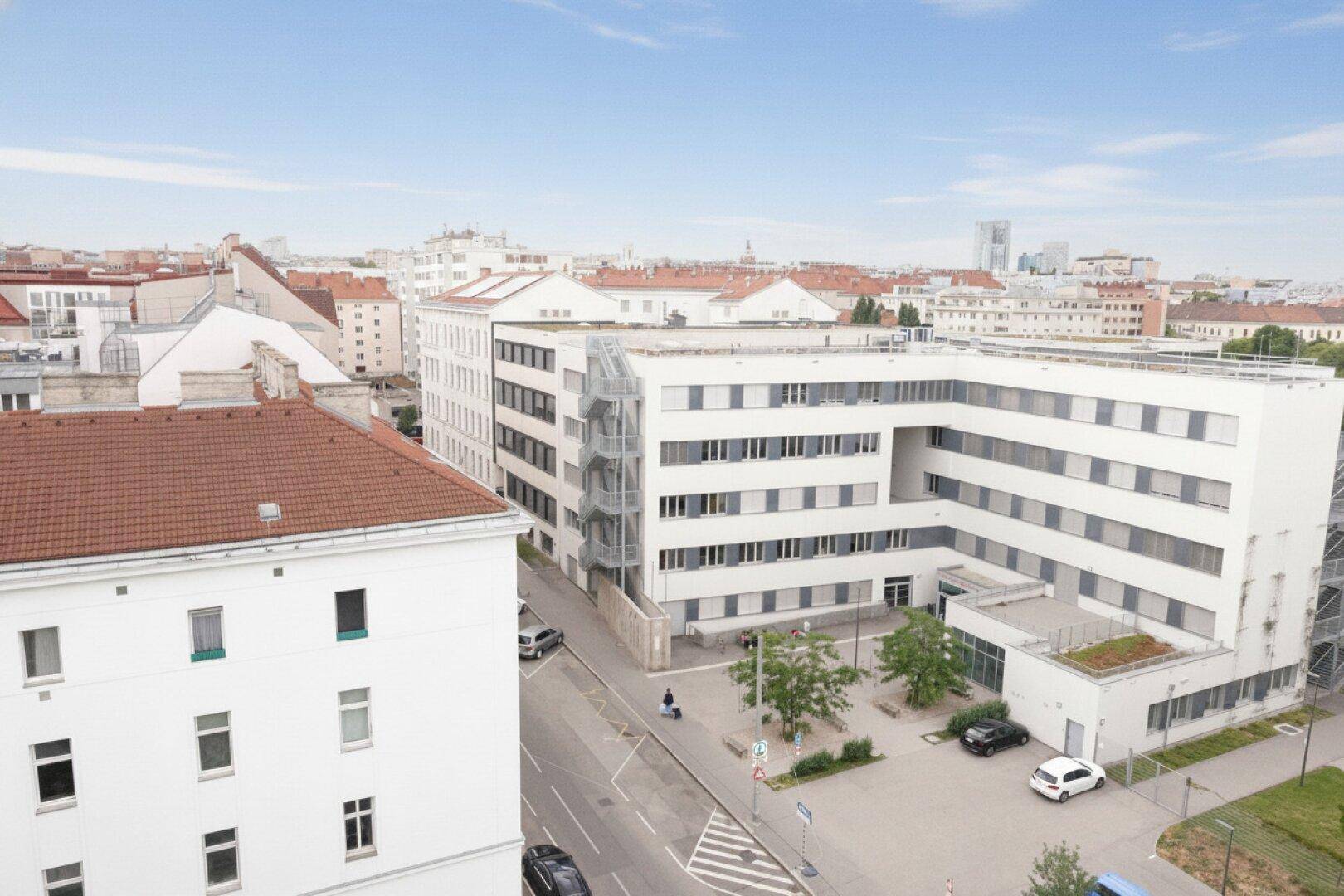 Luftaufnahme eines modernen Gebäudekomplexes in städtischer Umgebung mit umliegenden Dächern.