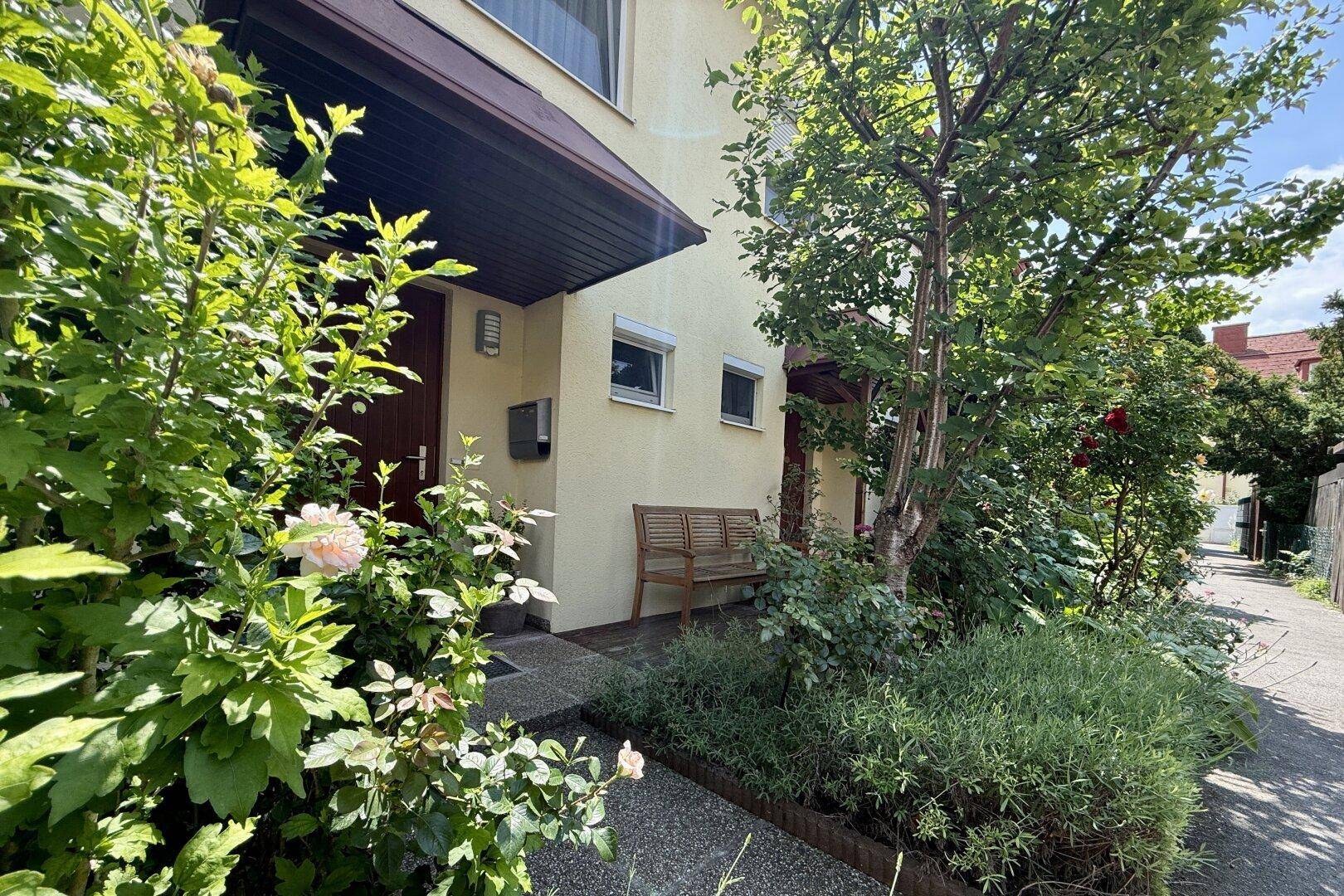Eingangsbereich des Hauses mit kleiner Gartenfläche, Holzbank und überdachtem Zugang.