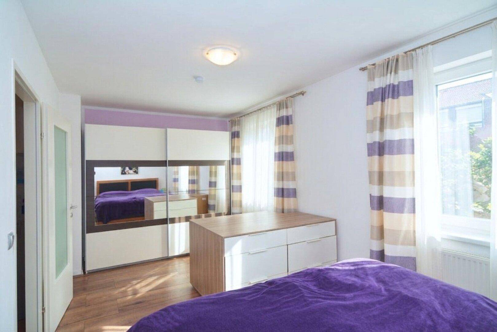 Schlafzimmer mit großem Spiegelschrank, Kommode und lila Bettwäsche.