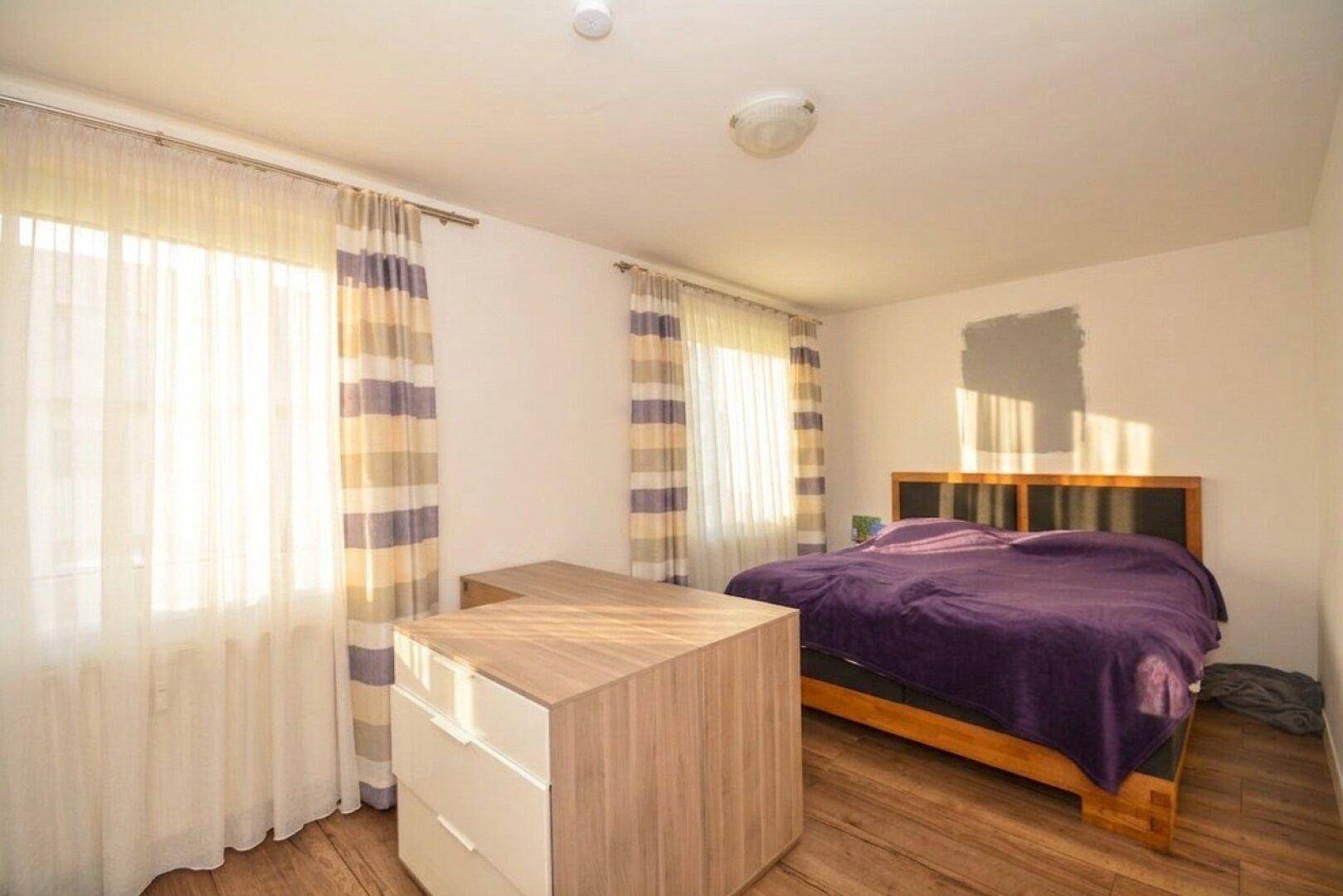 Helles Schlafzimmer mit Holzbett, lila Bettwäsche und zwei Fenstern mit Vorhängen.