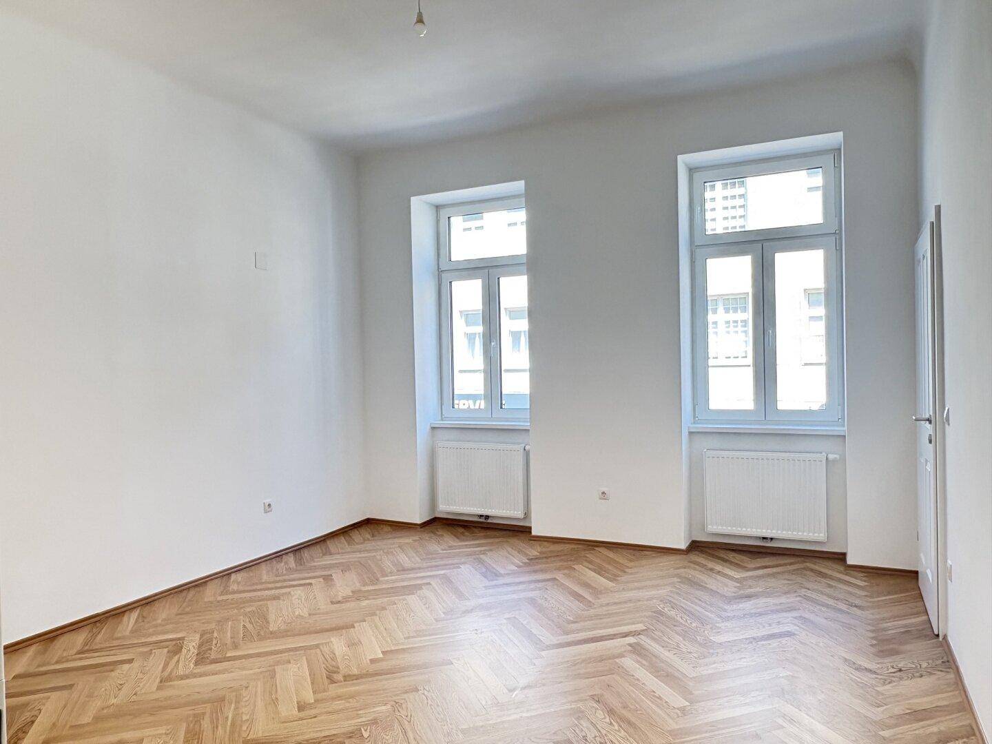 Leeres Zimmer mit Fischgrätparkett, hohen Fenstern und weiß gestrichenen Wänden.