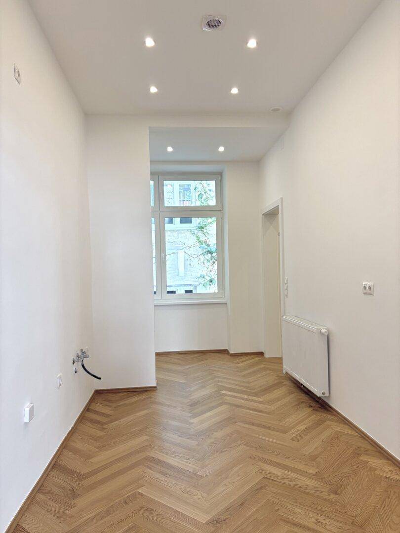 Leerer Küchenbereich mit Fischgrätparkett, Einbaustrahlern und einem Fenster zum Innenhof.