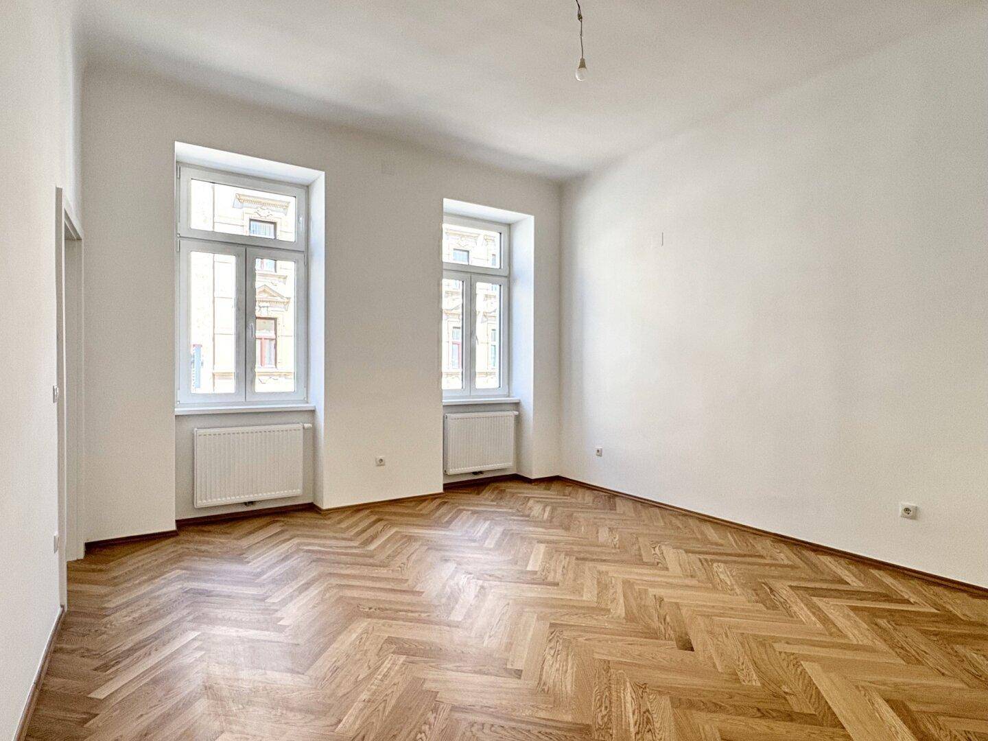 Geräumiges, leeres Schlafzimmer mit Fischgrätparkett und zwei großen Fenstern.