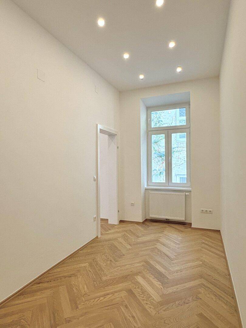 Leeres Zimmer mit Fischgrätparkett, Einbaustrahlern und einem Fenster zum Innenhof.