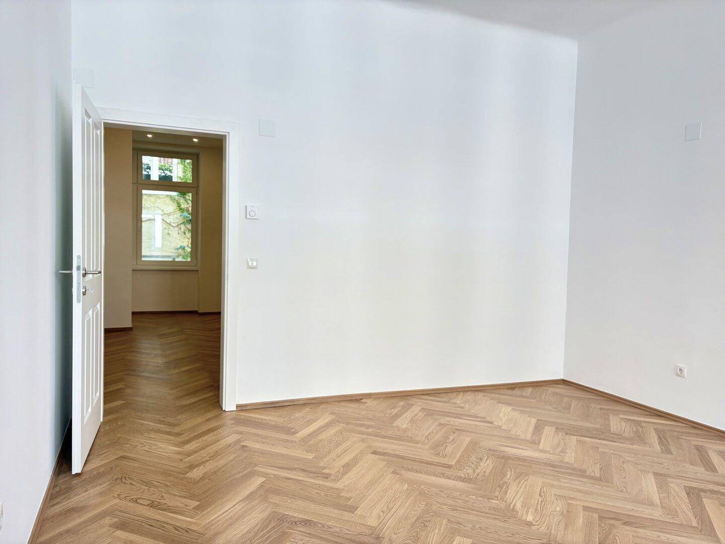 Leerer Raum mit Fischgrätparkett und einer Tür, die zu einem weiteren Zimmer führt.